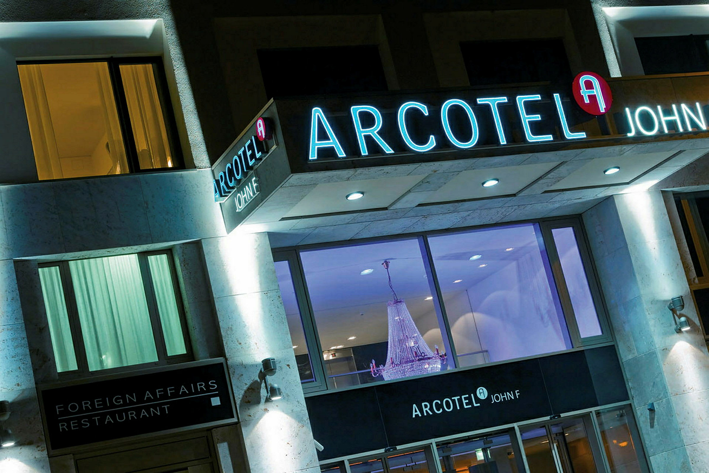 Billede af en hotelfacade med lysskilt, der viser navnet Arcotel John F, samt en restaurant kaldet Foreign Affairs. En stor lysekrone ses gennem de store vinduer. (AI-genereret tekst)