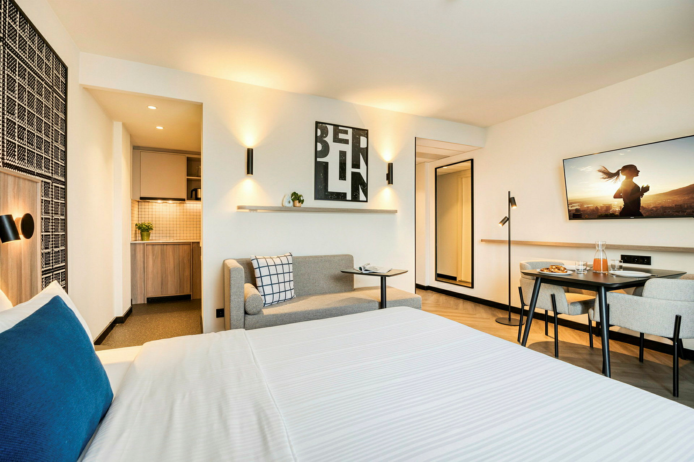 Interiør af et moderne hotelværelse med seng, lille køkkenområde og siddegruppe. (AI-genereret tekst)