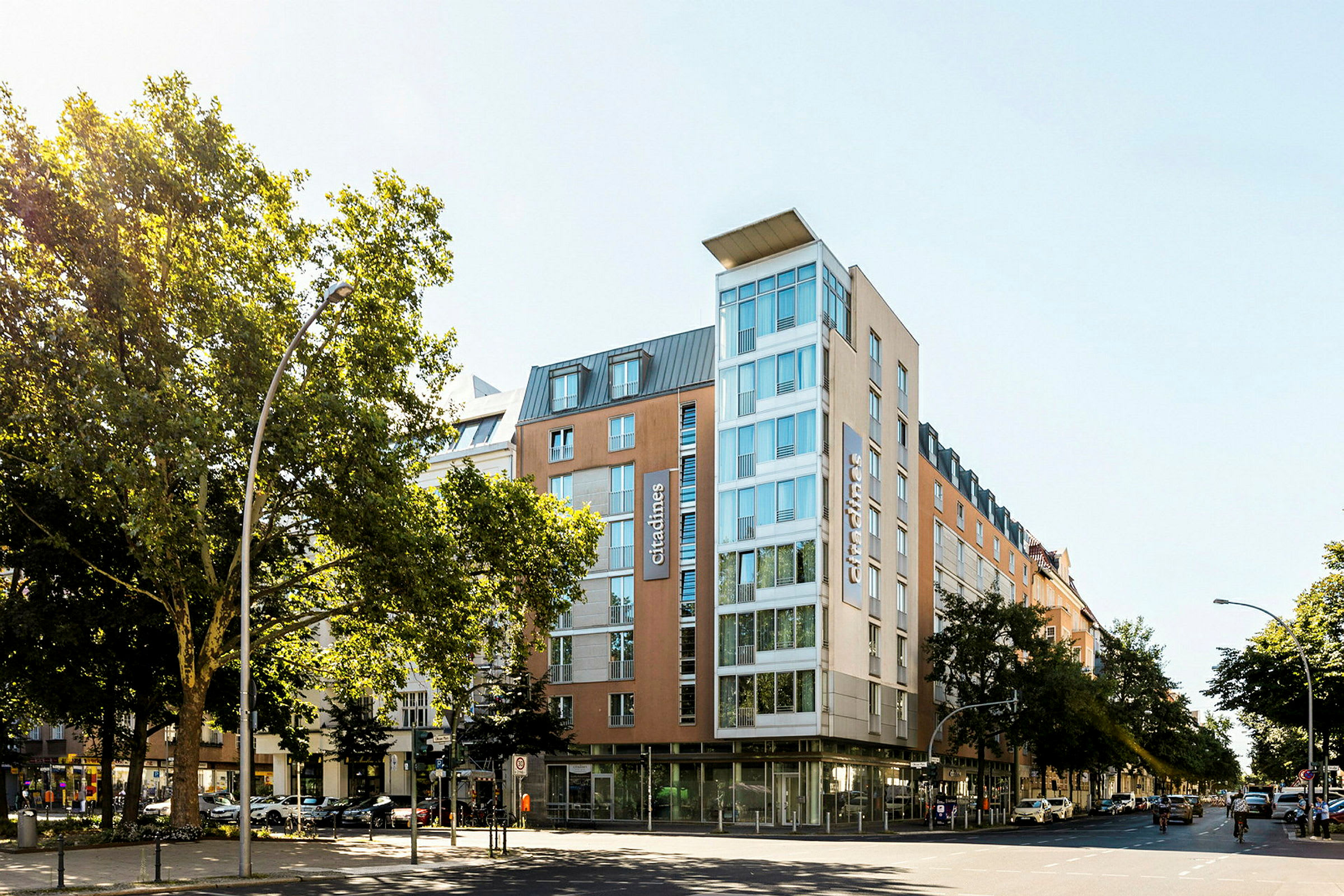 Et moderne hotel med glasfacade omgivet af træer og bymiljø. (AI-genereret tekst)