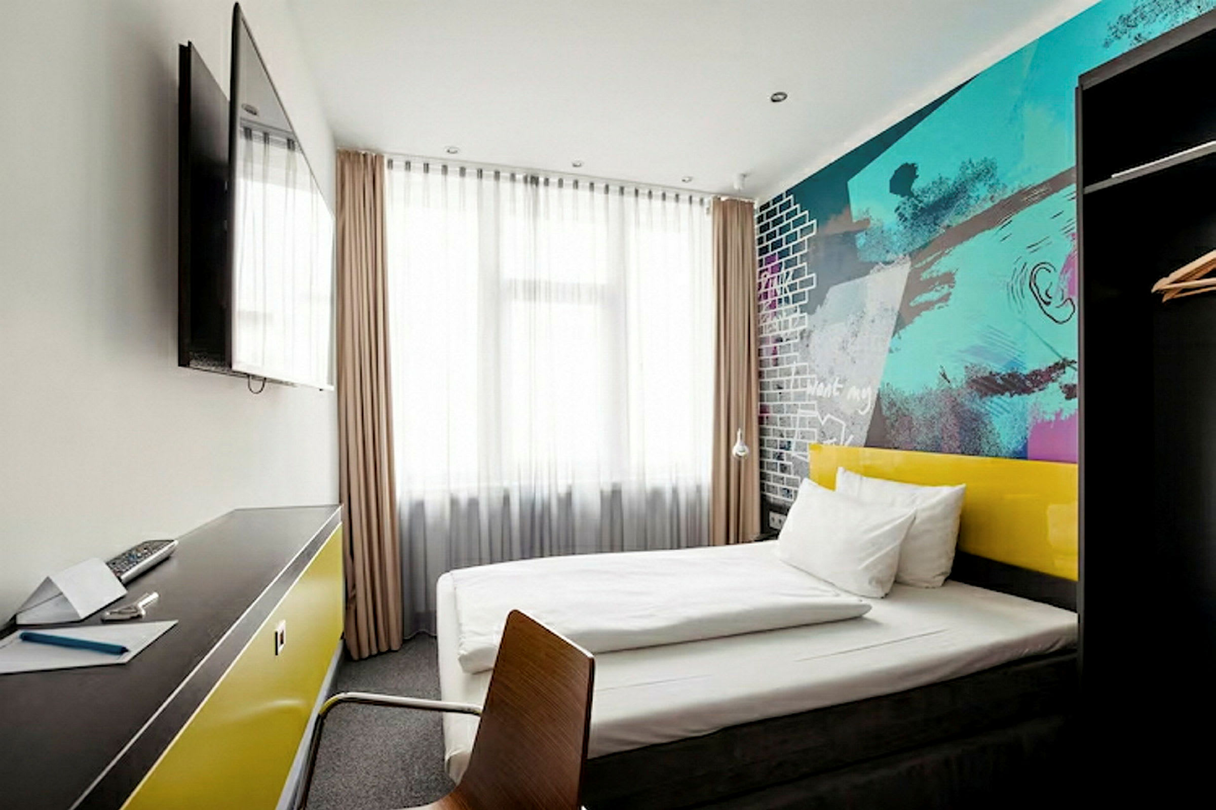 Et moderne hotelværelse med en seng, farverige vægge og et skrivebord med et fjernsyn på væggen. (AI-genereret tekst)