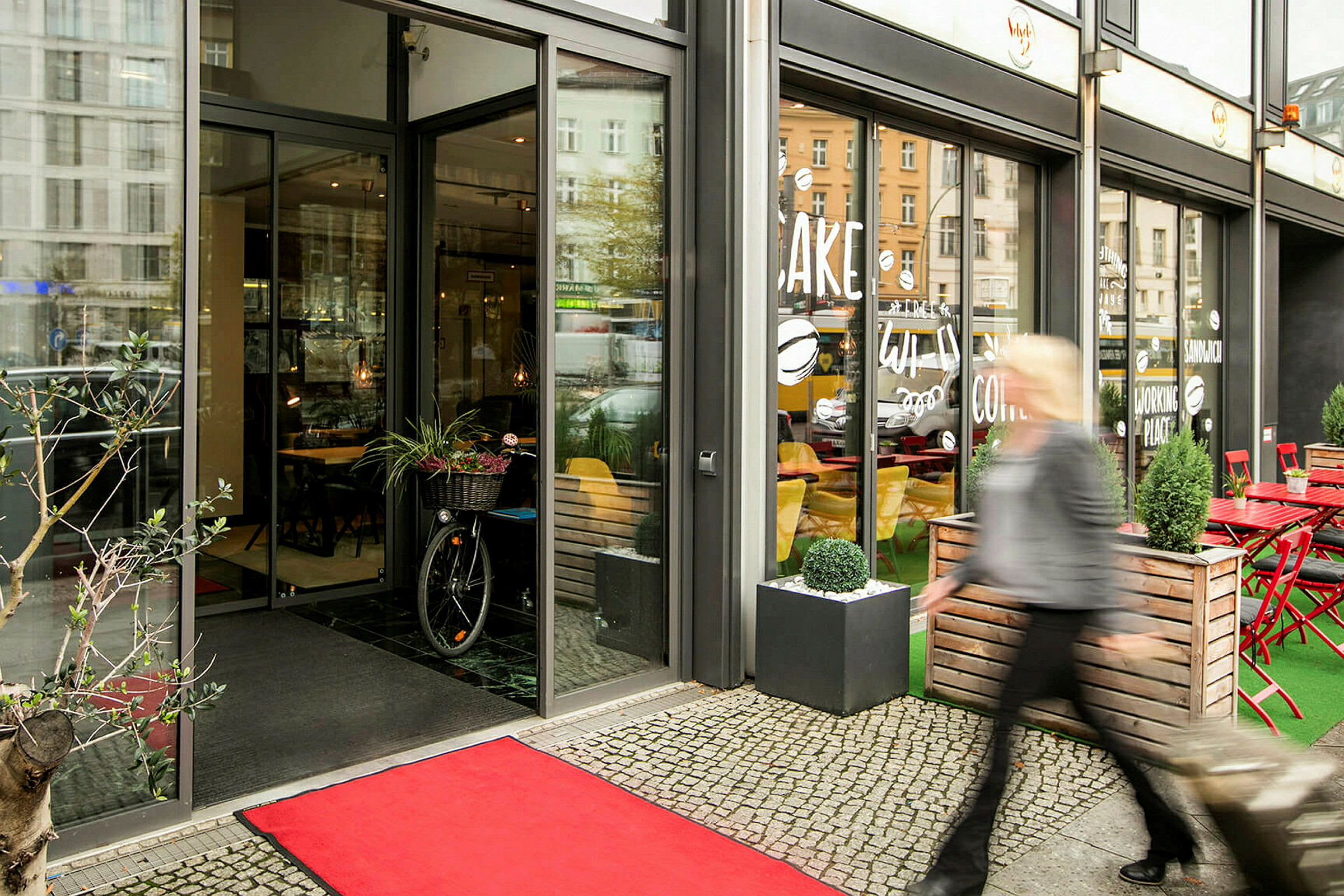 Indgang til en café med glasfacade, rødt tæppe på jorden og en person, der går forbi udenfor. (AI-genereret tekst)
