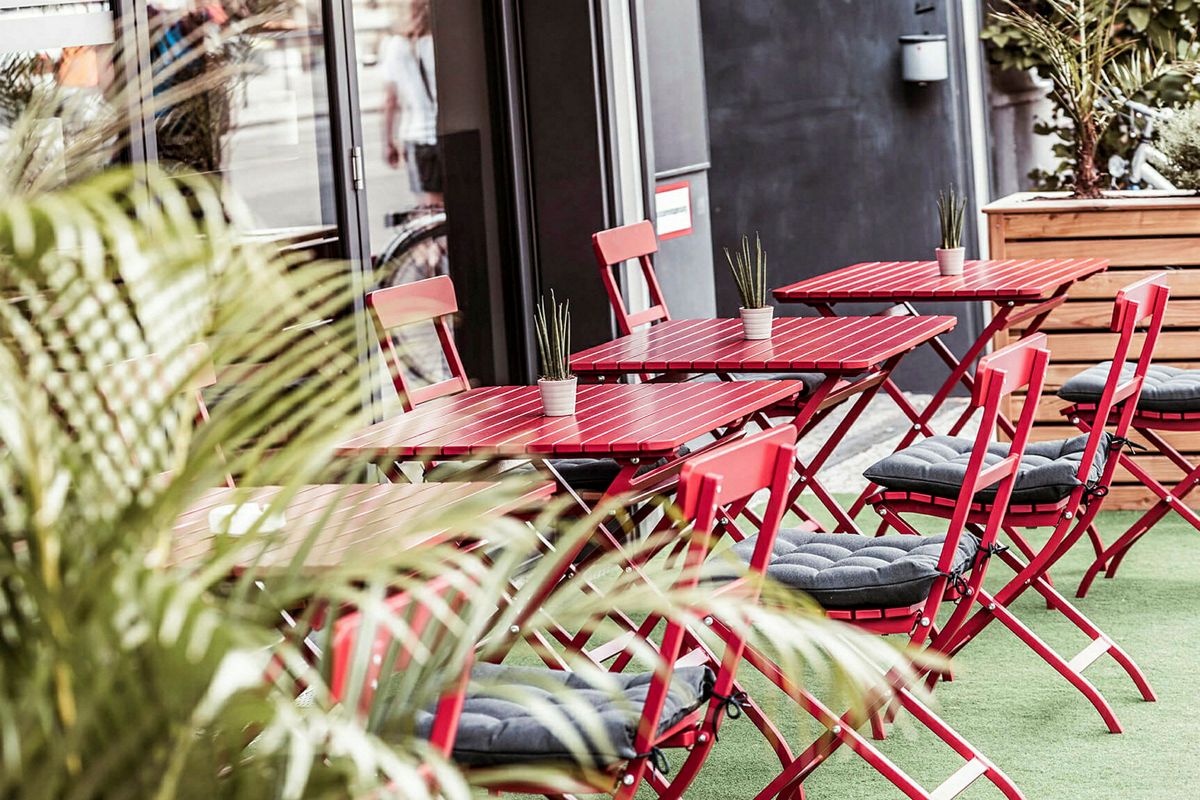 Billede af en udendørs café med røde borde og stole samt små plantepynt. (AI-genereret tekst)