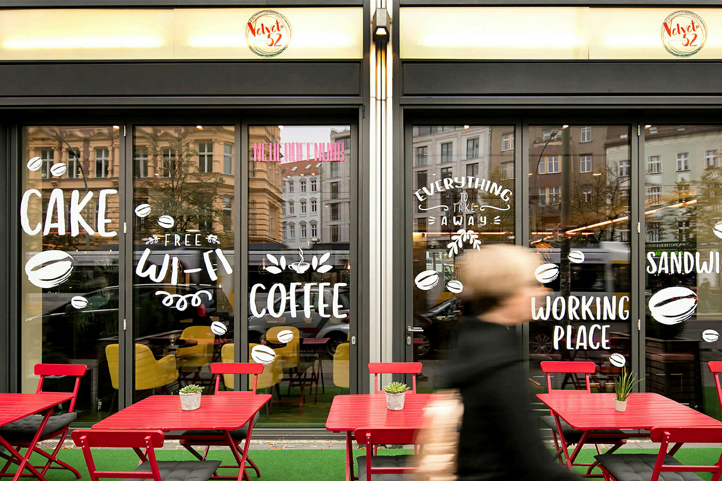 Forsiden af en café med store glasvinduer dekoreret med tekst og symboler. Udenfor står røde borde og stole. (AI-genereret tekst)