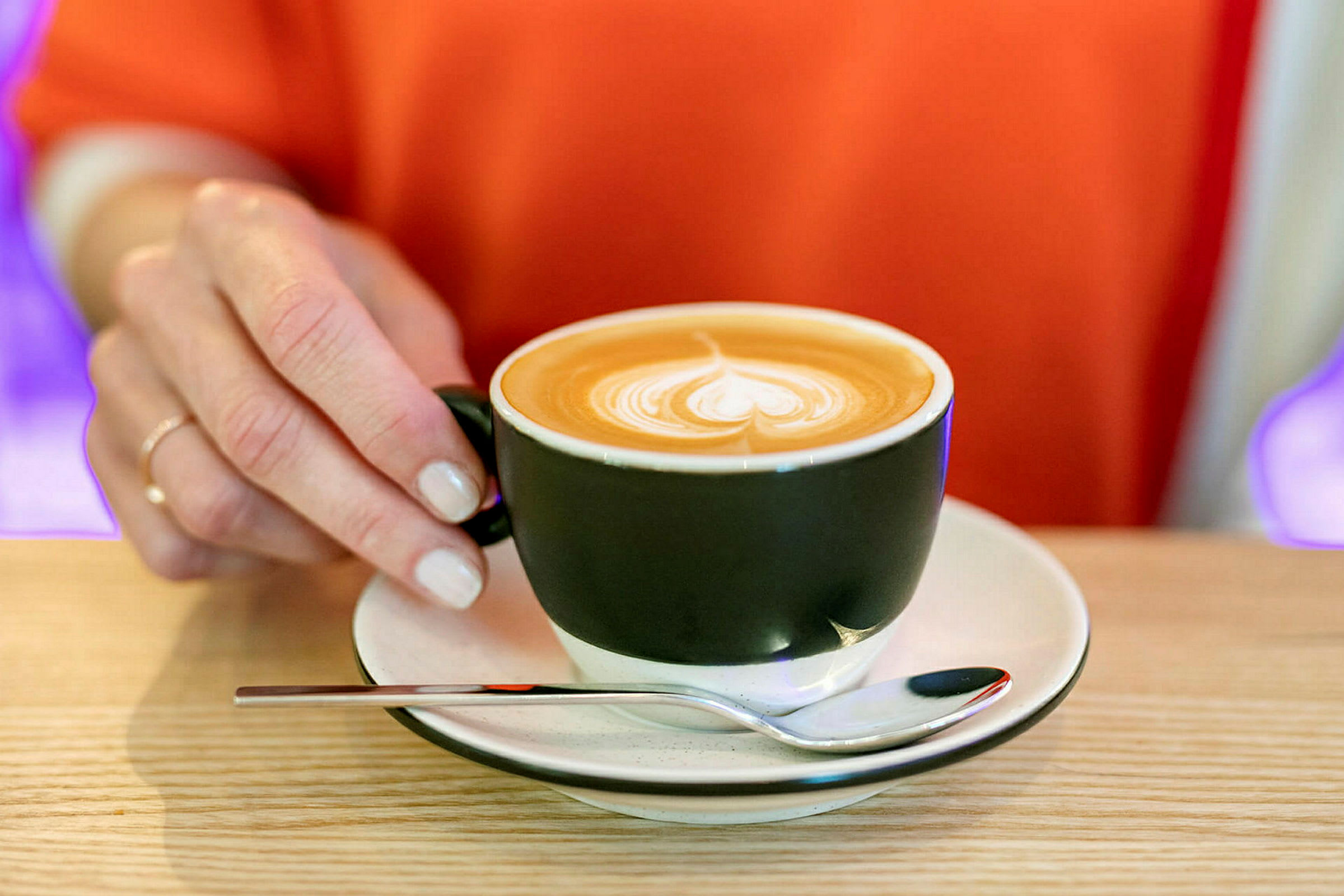 En hånd holder på et sort kaffekopøre med en underkop og en ske på et træbord. Cappuccino med latte art på toppen. (AI-genereret tekst)
