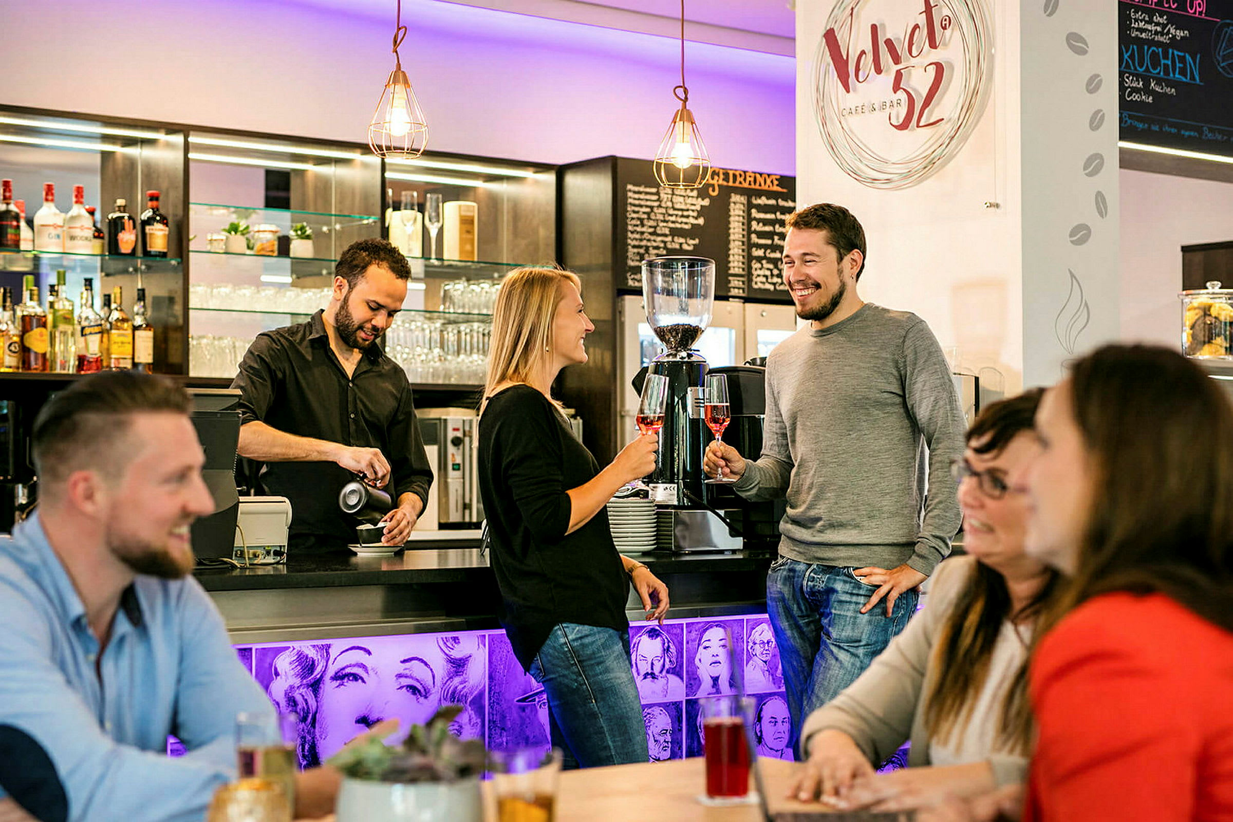En barista hælder kaffe ved en bardisk, hvor folk står og sidder og nyder deres drikke. (AI-genereret tekst)