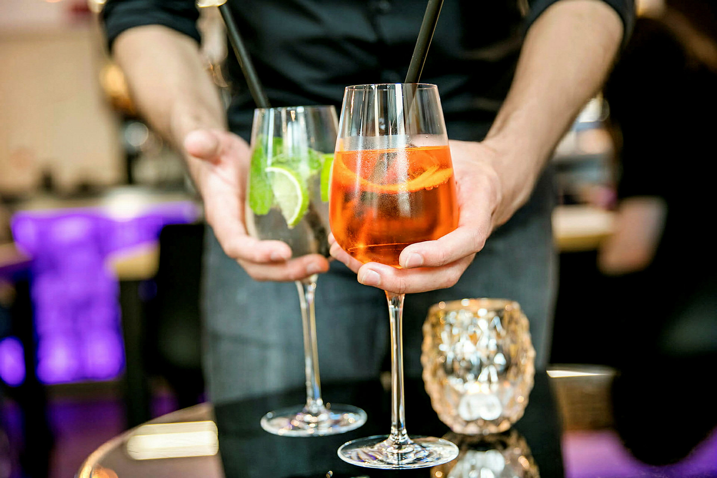 To drinks holdes frem af en person, et glas indeholder en orange cocktail og det andet en klar cocktail med lime og mynte. (AI-genereret tekst)