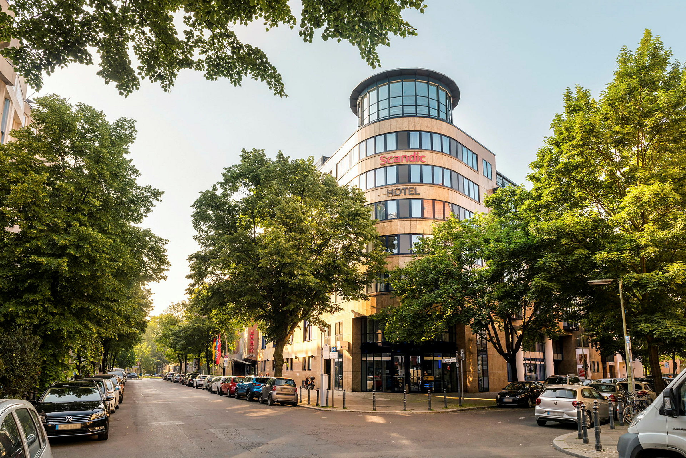En byudsigt med facaden af et Scandic-hotel omgivet af træer og parkerede biler. (AI-genereret tekst)