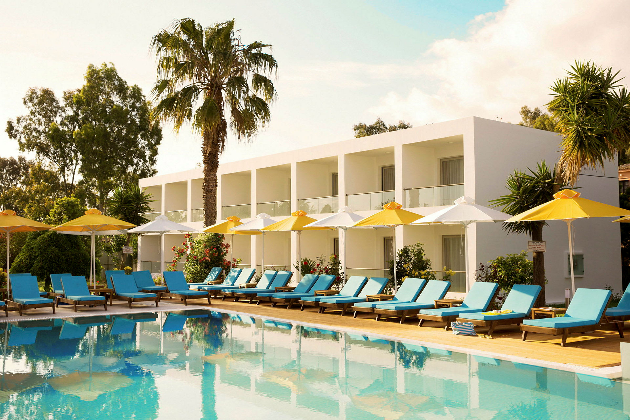 Et hotel med hvide bygninger og altaner i en grøn omgivelser, foran en pool med liggestole og parasoller. (AI-genereret tekst)