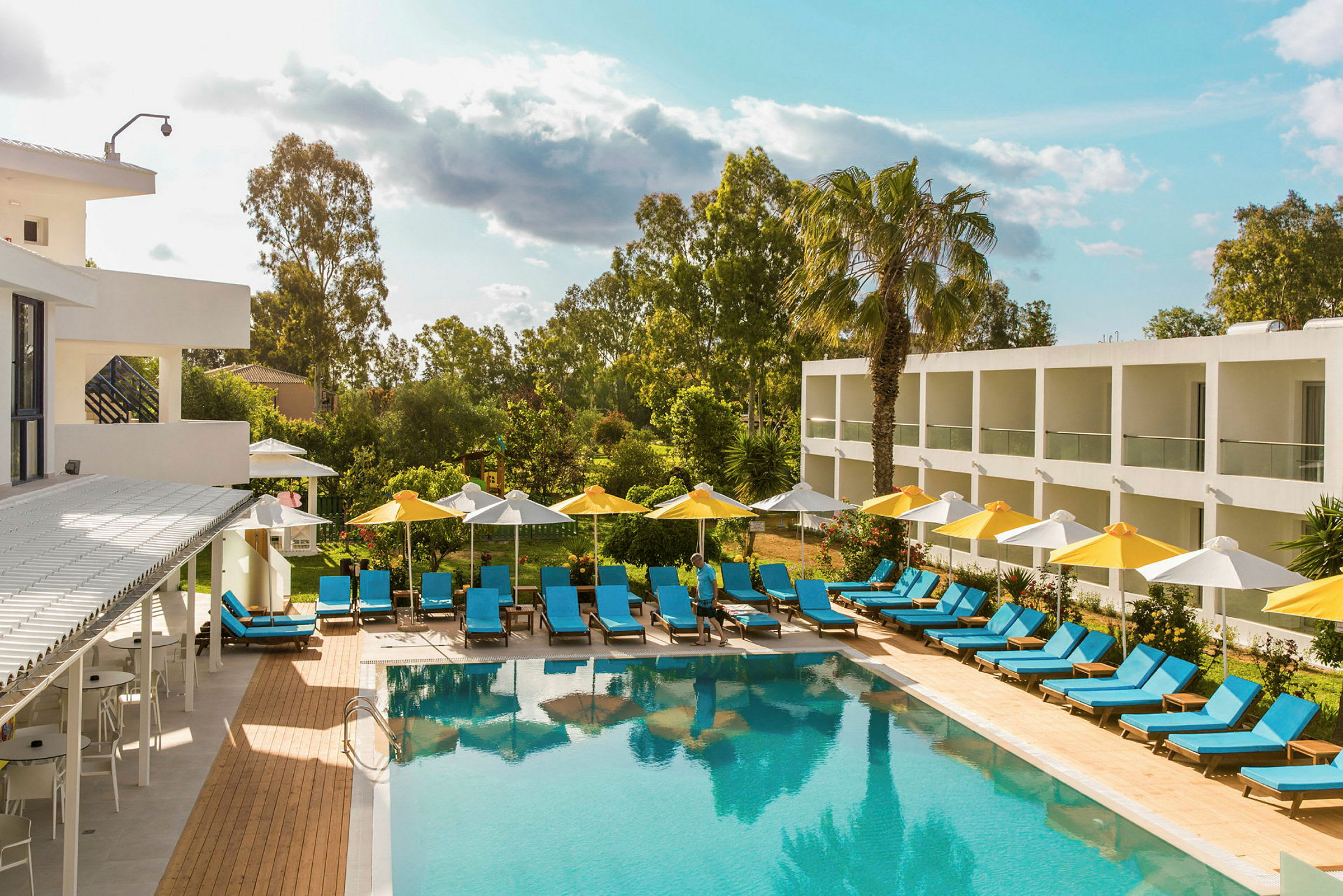Et hotelområde med en udendørs pool omgivet af liggestole og parasoller. Bygninger og vegetation er synlige i baggrunden. (AI-genereret tekst)