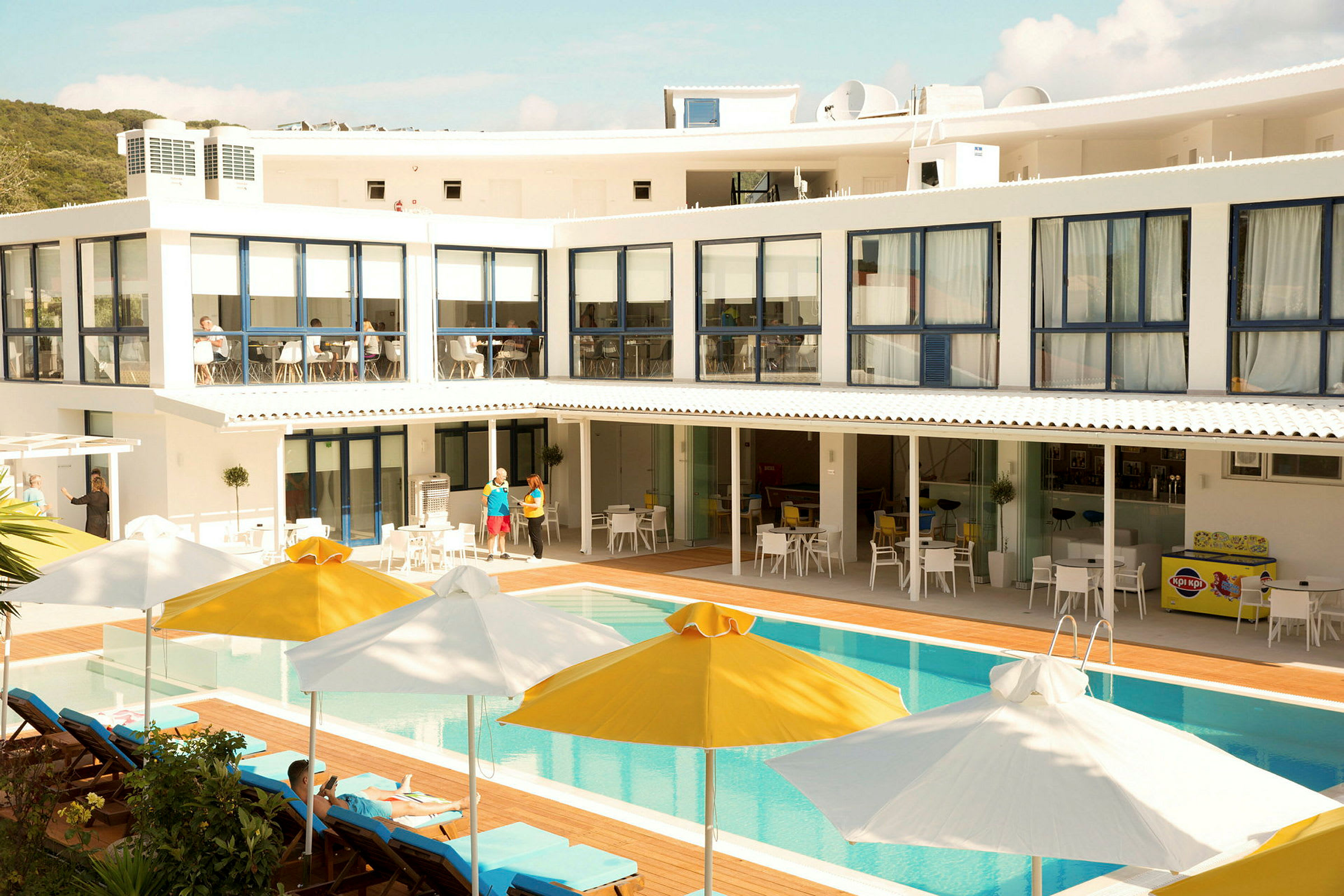 Et billede af en hotelbygning med swimmingpool og liggestole, samt parasoller i gul og hvid. (AI-genereret tekst)