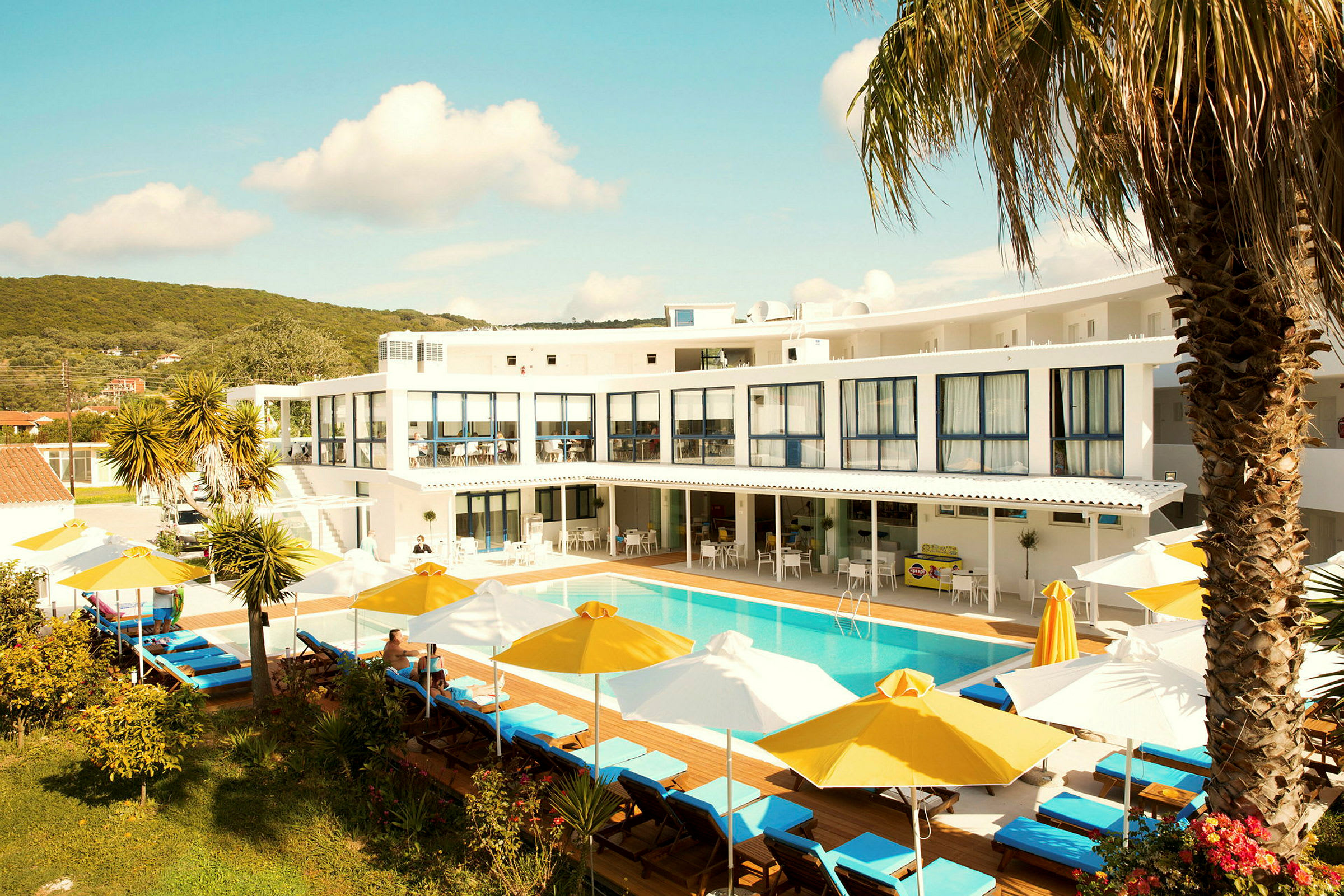 Et hotel med hvid facade, en udendørs pool omgivet af liggestole med gule og hvide parasoller samt beplantning i forgrunden. (AI-genereret tekst)