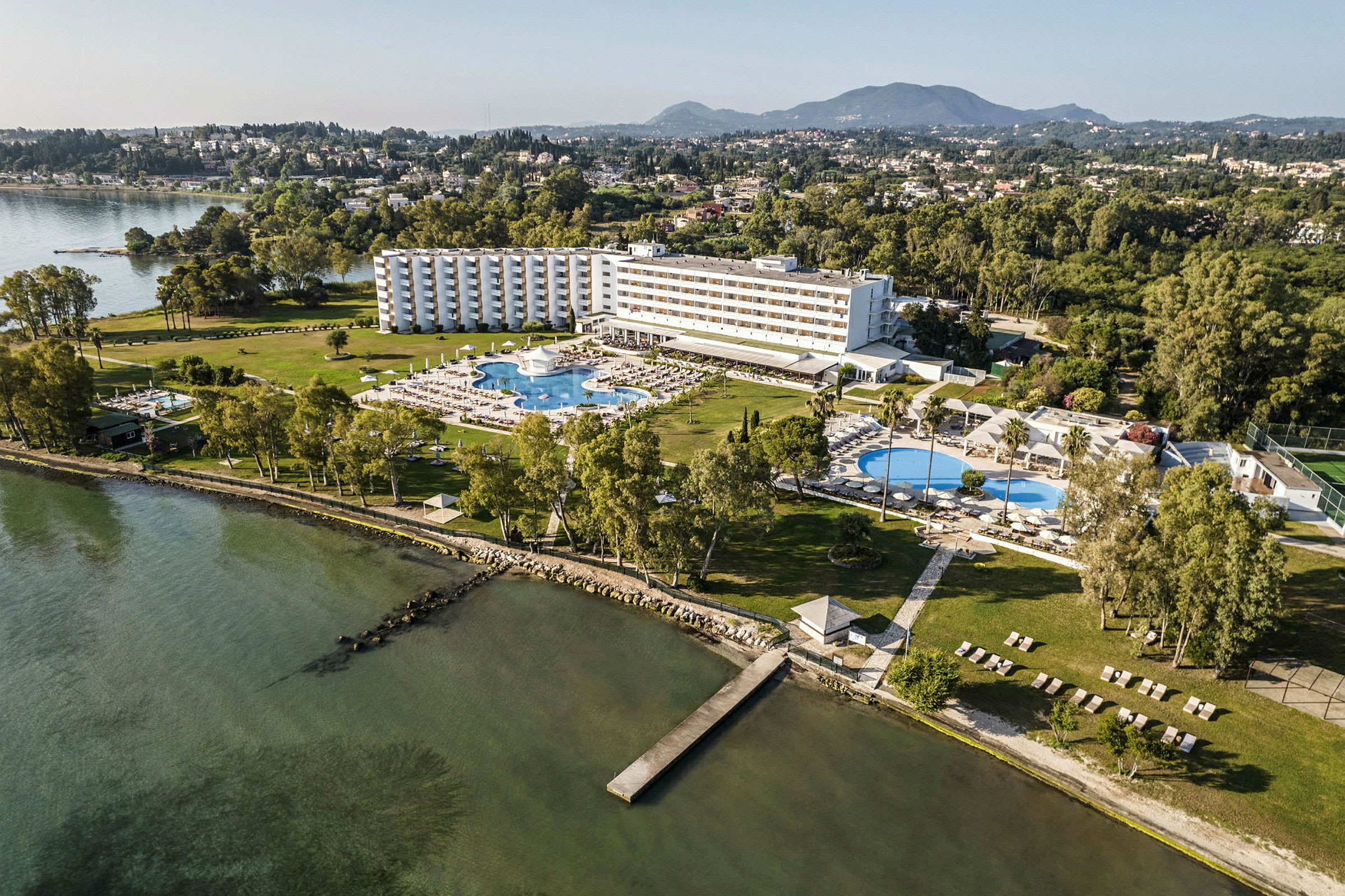 Luftfoto af et hotel med omkringliggende haver, pools og tæt på en kystlinje. (AI-genereret tekst)
