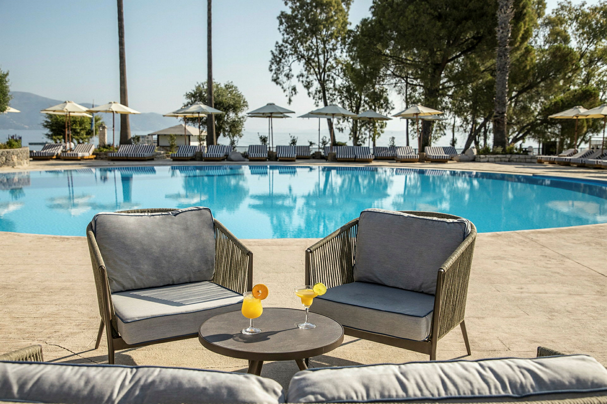 To lænestole med bord og cocktails ved en swimmingpool med solvogne i baggrunden. (AI-genereret tekst)