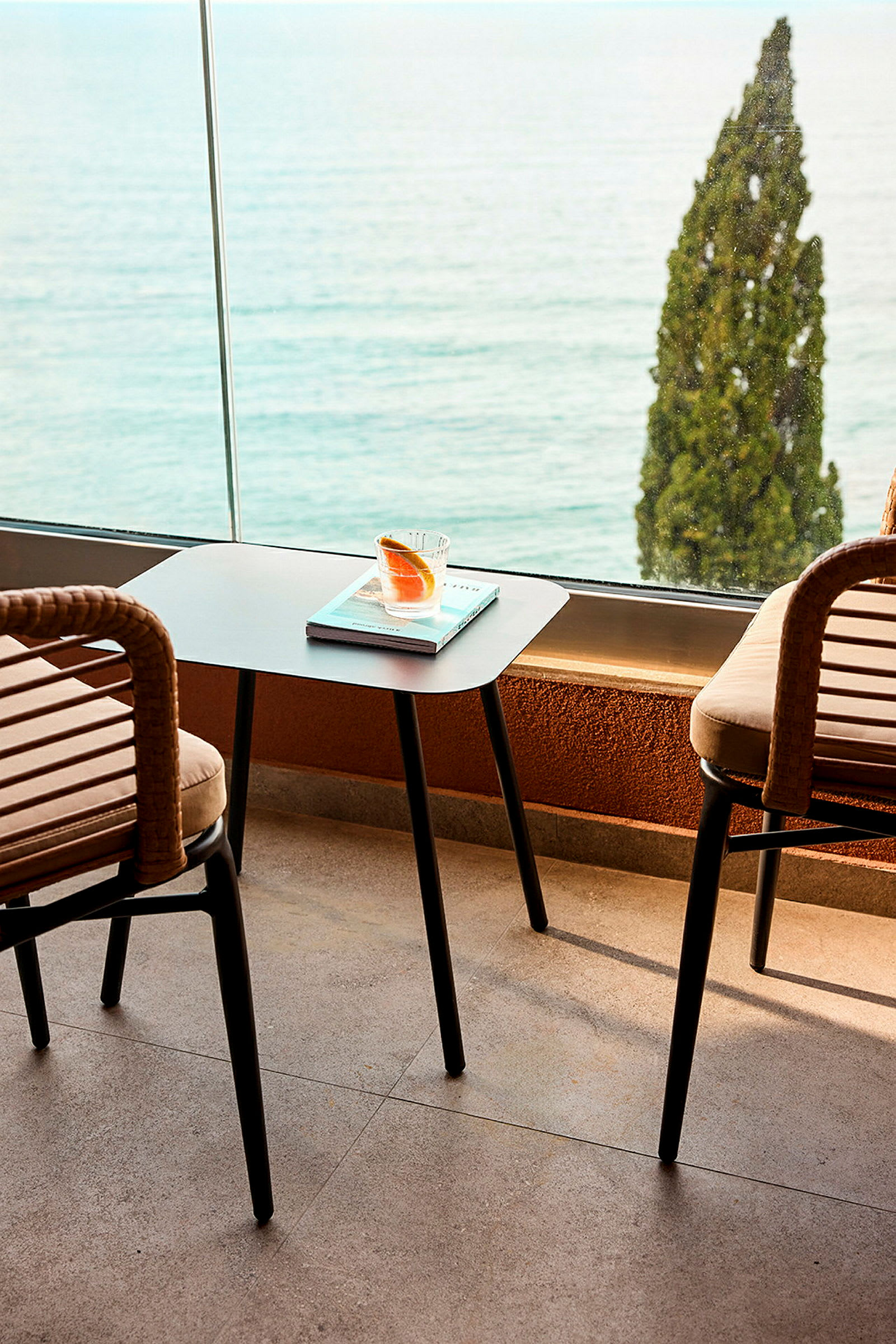 To stole og et bord med et glas og en bog står på en terrasse med udsigt over havet og et træ. (AI-genereret tekst)