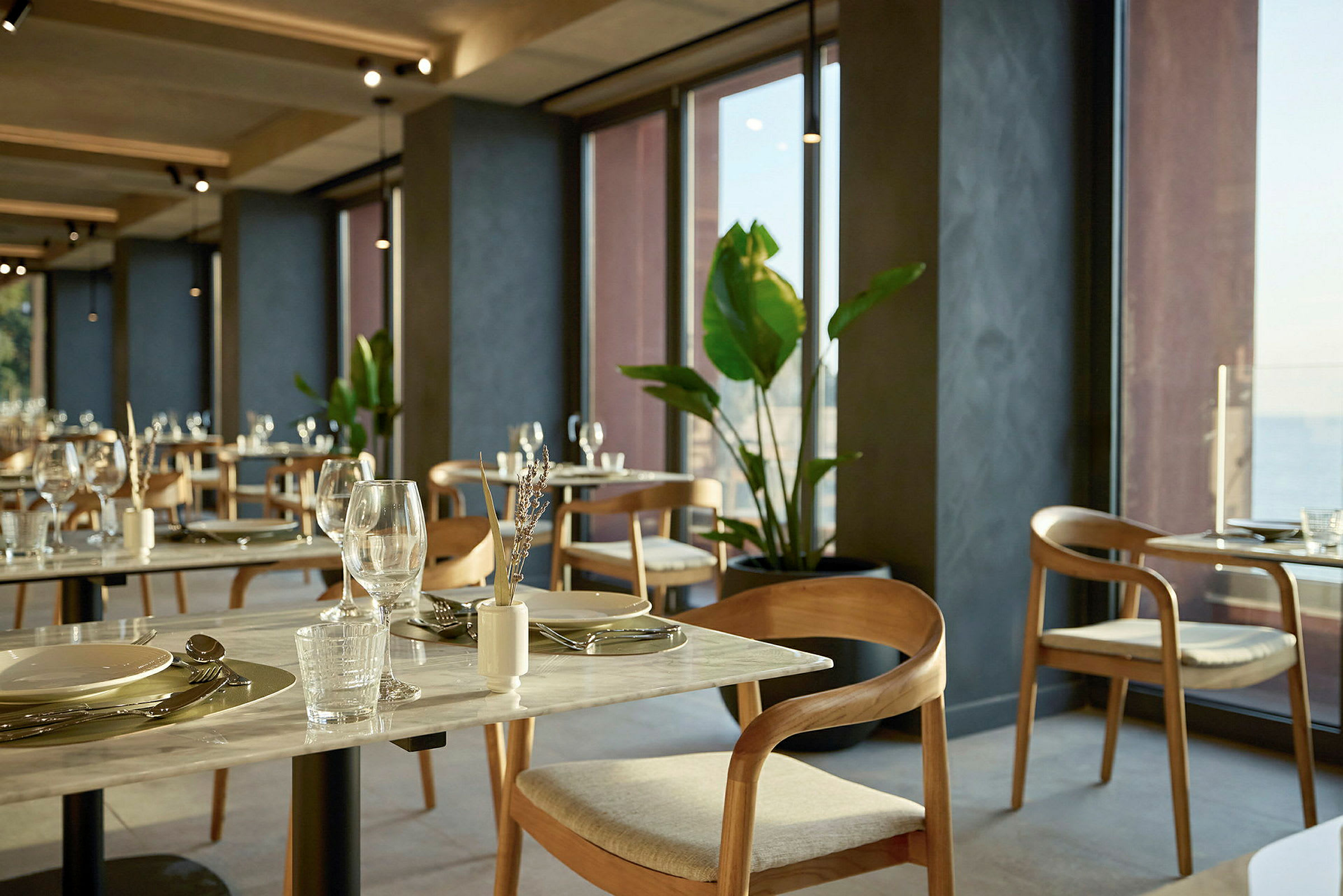 En restaurant med borde og stole ved store vinduer, dekoreret med planter. (AI-genereret tekst)