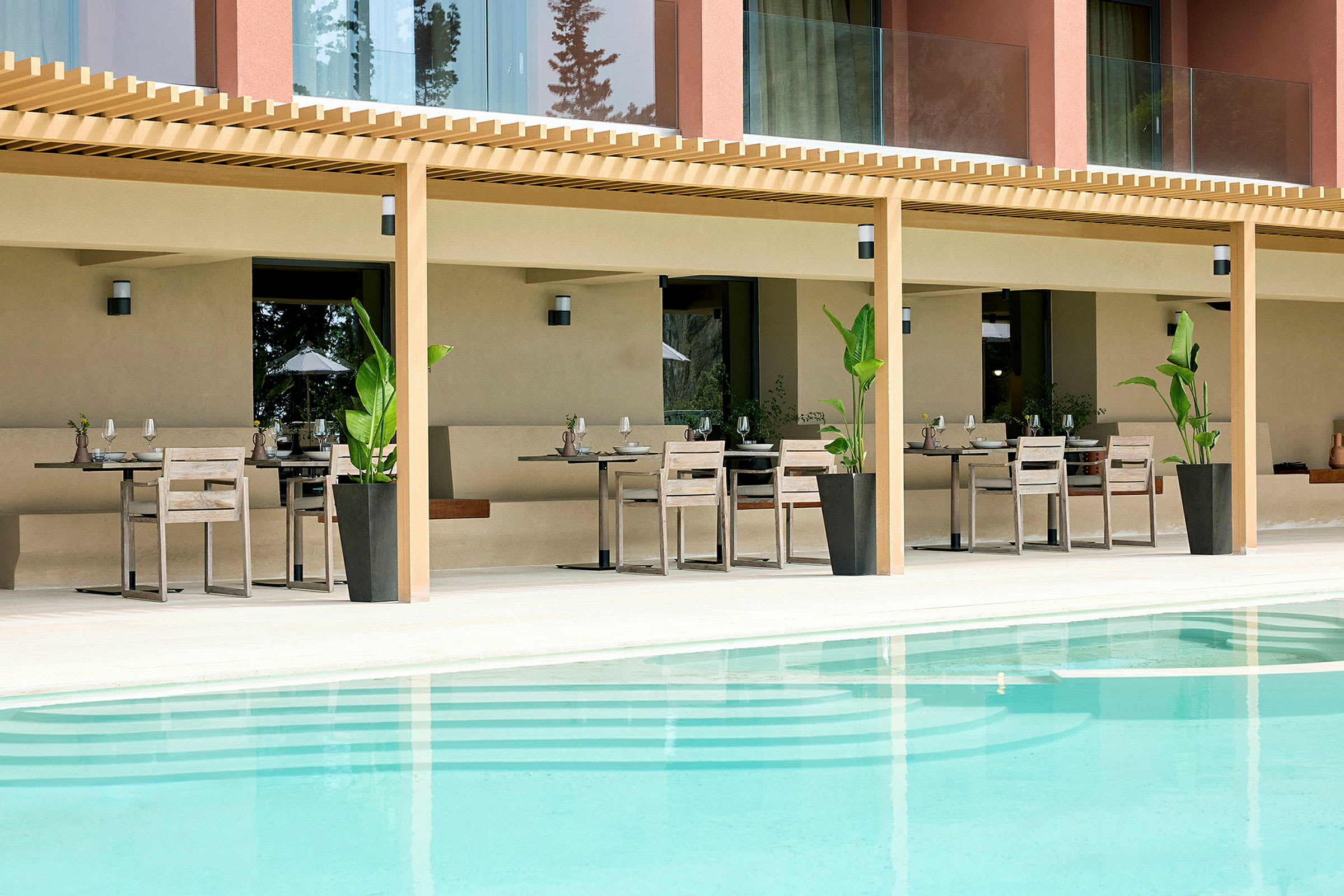 En udendørs servering med borde og stole under en pergola foran en swimmingpool. (AI-genereret tekst)