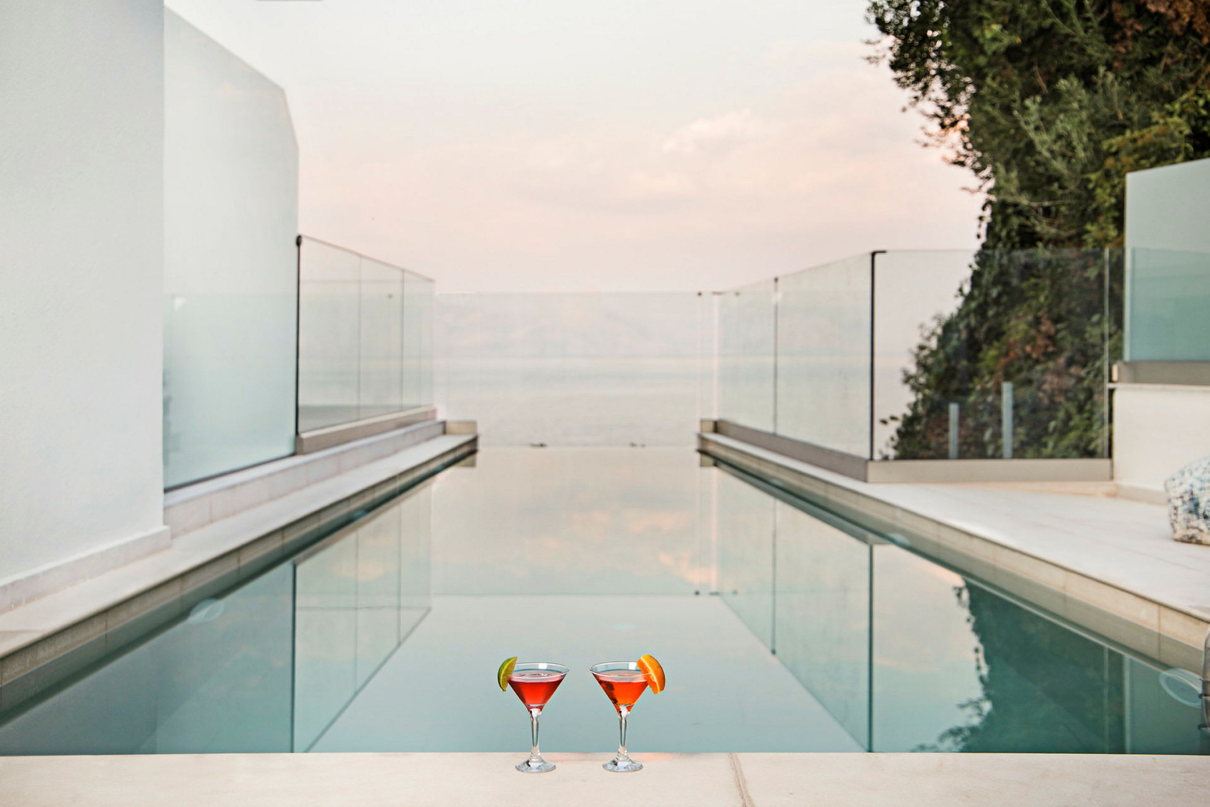 To glas med orange-rød drik står ved kanten af en pool omgivet af glasrækværk og med udsigt over havet. (AI-genereret tekst)
