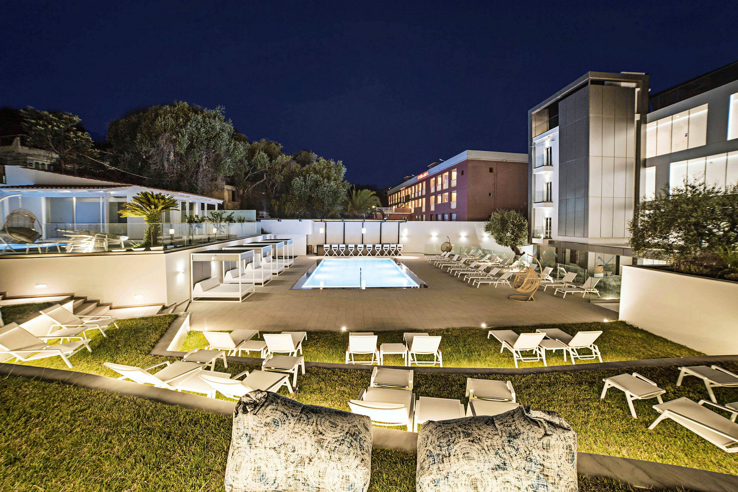 Et oplyst hotelområde om aftenen med en pool omgivet af solsenge og moderne bygninger i baggrunden. (AI-genereret tekst)