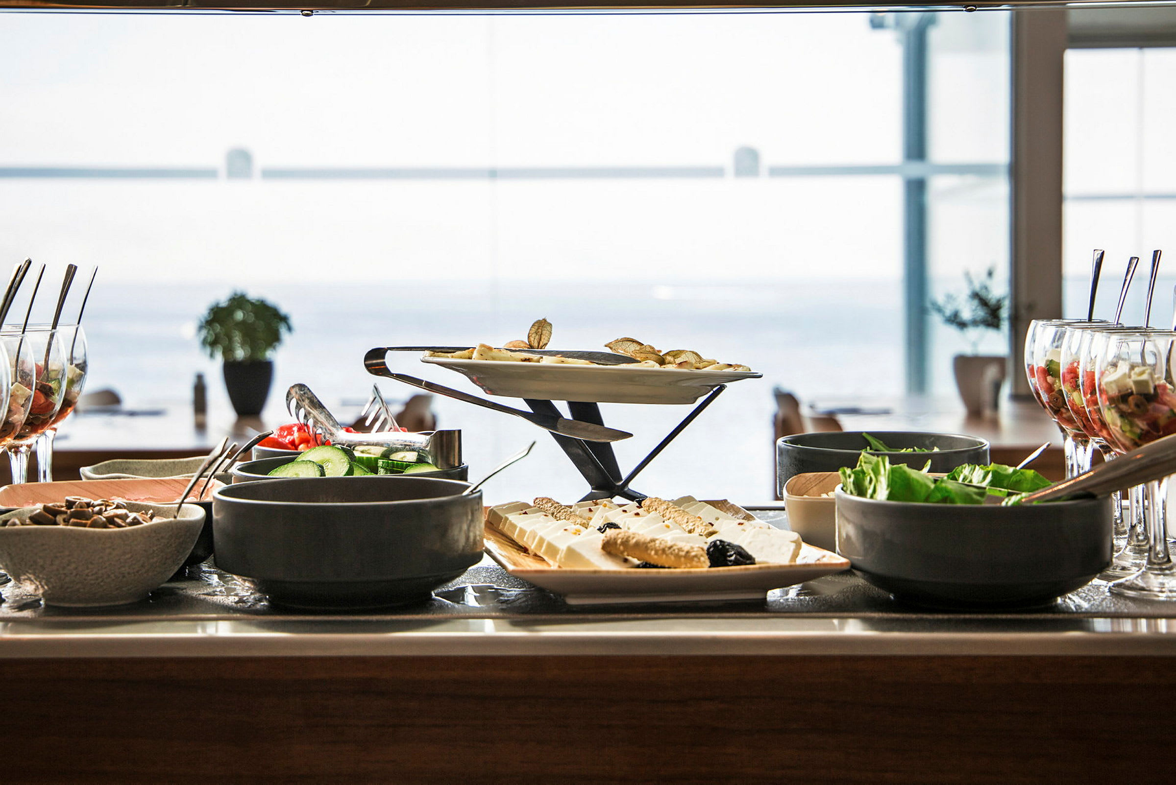 Buffet med forskellige retter, salater og desserter på et bord med havudsigt i baggrunden. (AI-genereret tekst)