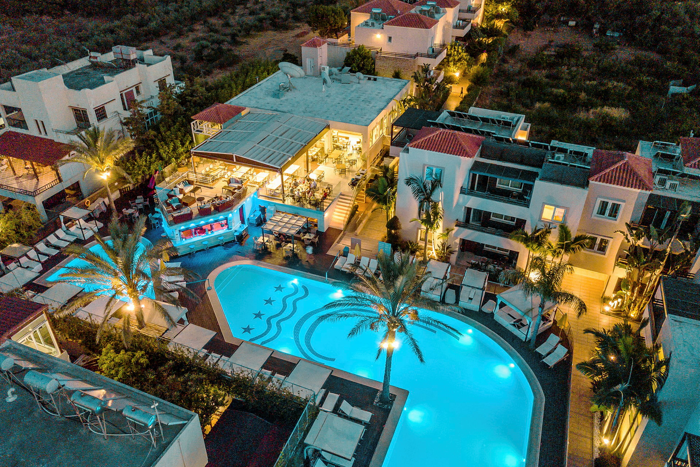 Luftfoto af et hotelområde med oplyst pool og bygninger omkring, en palmelignende plante står ved poolen. (AI-genereret tekst)