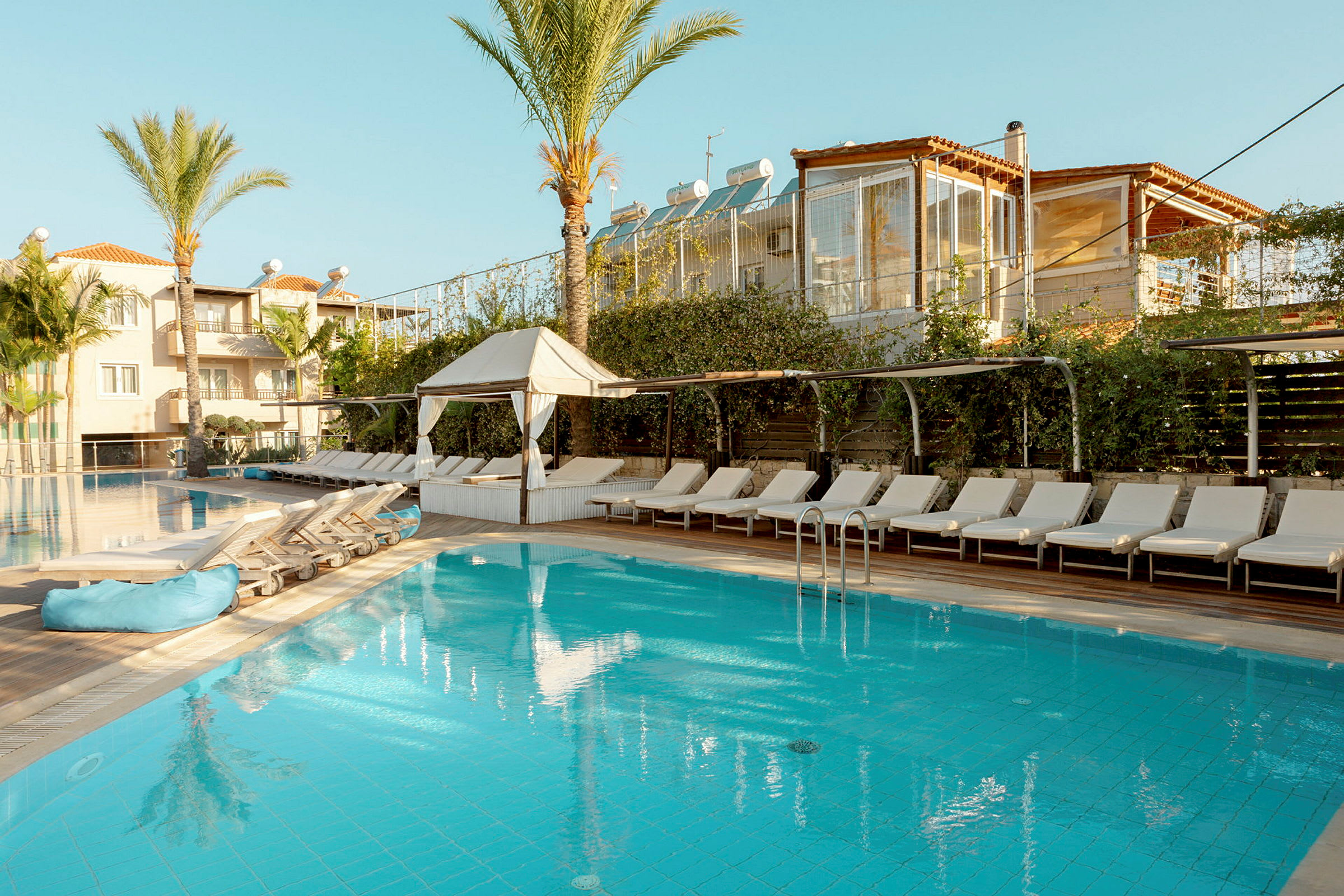 Billede af en pool omgivet af liggestole, med et hotel i baggrunden. (AI-genereret tekst)