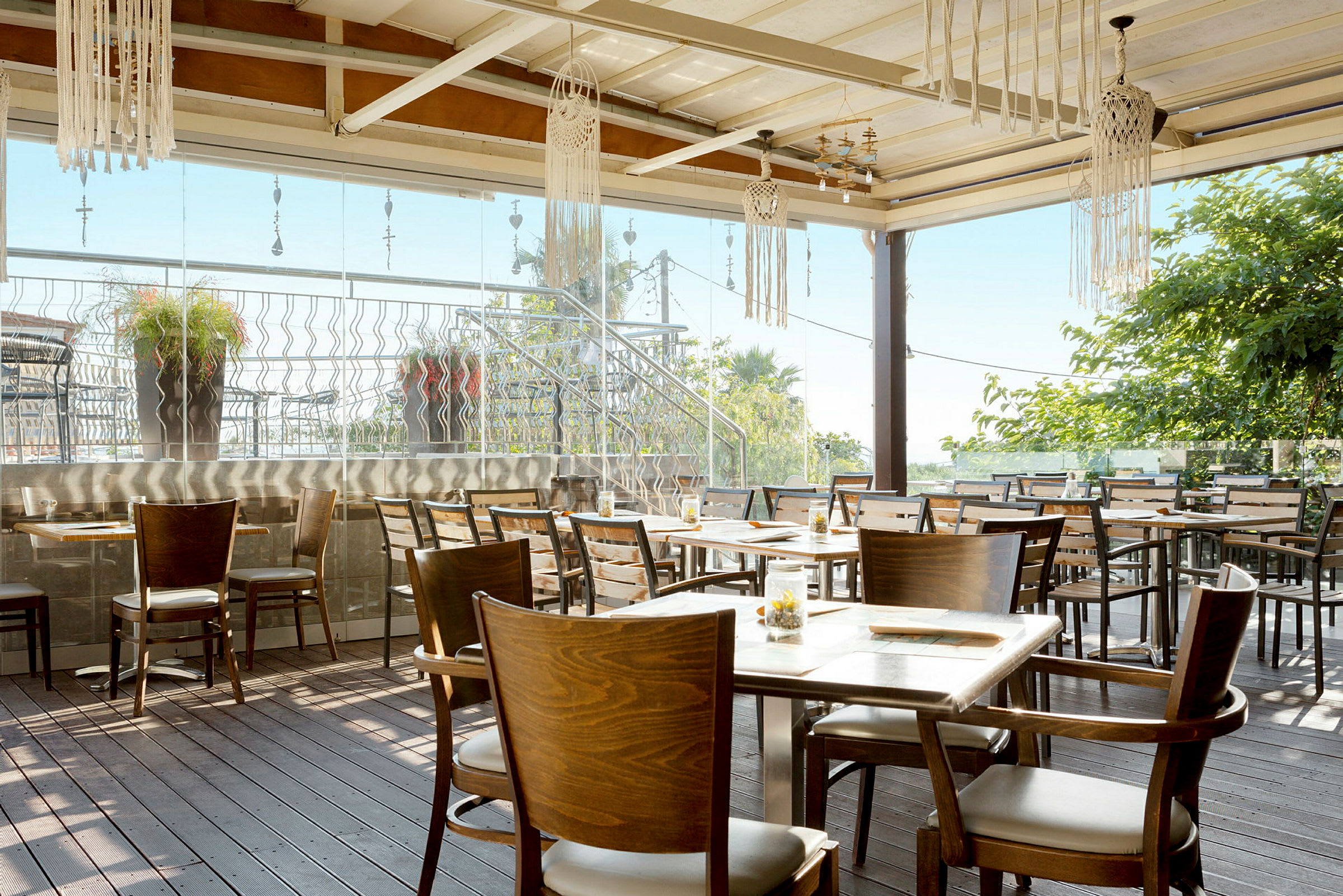 Billede af en udendørs terrasse med træborde og stole. Der hænger dekorationer fra loftet, og der er beplantning i baggrunden. (AI-genereret tekst)