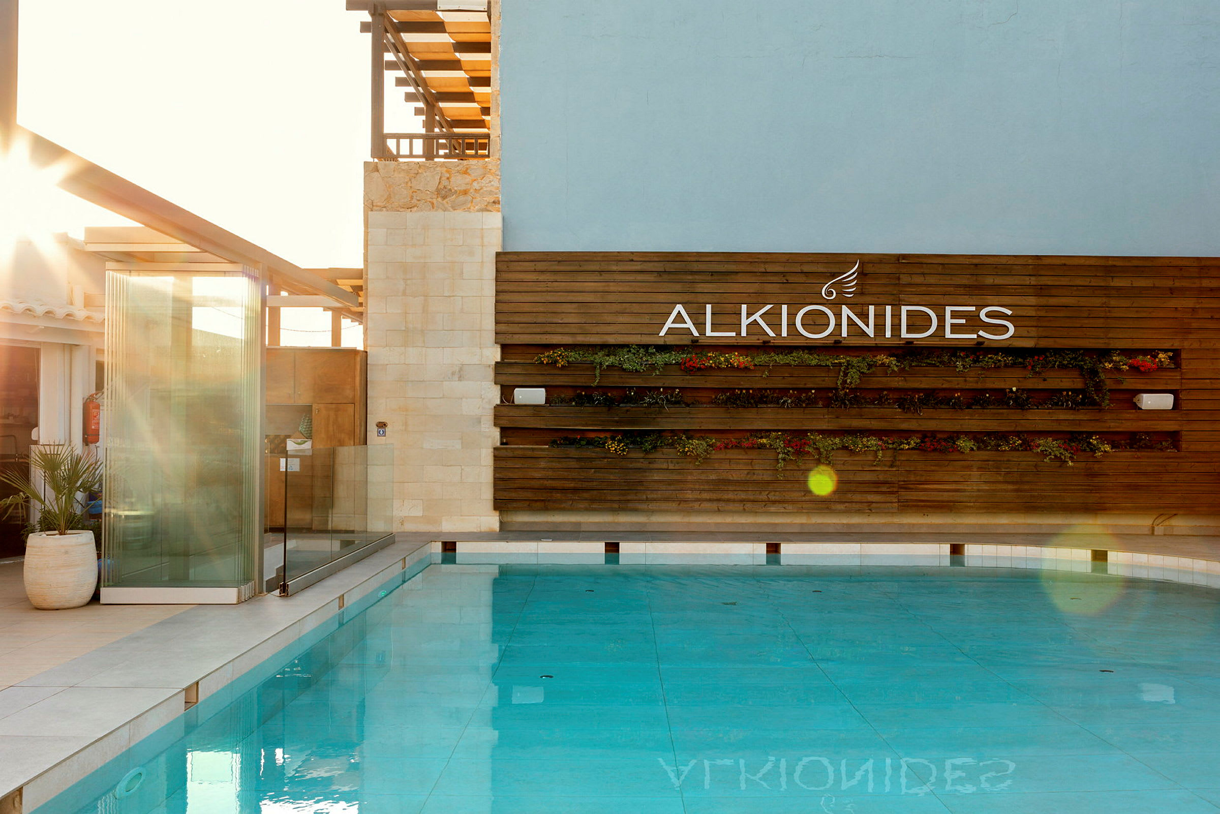 Billede af en pool med en trævæg i baggrunden, hvor der står "ALKIONIDES" samt dekorationer med planter. (AI-genereret tekst)