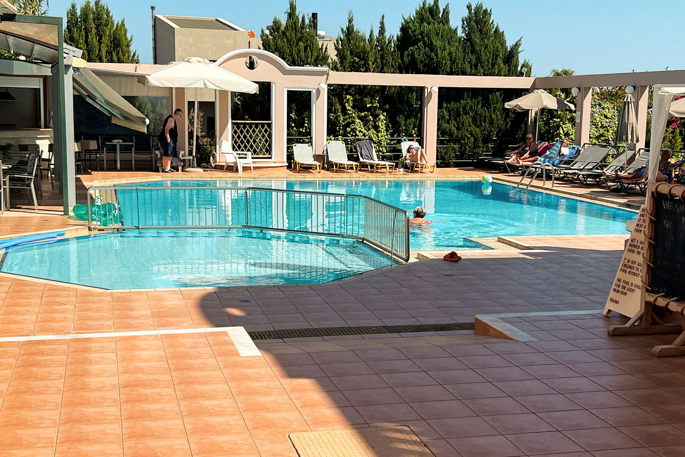 Udendørs swimmingpool med liggestole og parasoller ved siden, samt personer der svømmer og soler sig. (AI-genereret tekst)