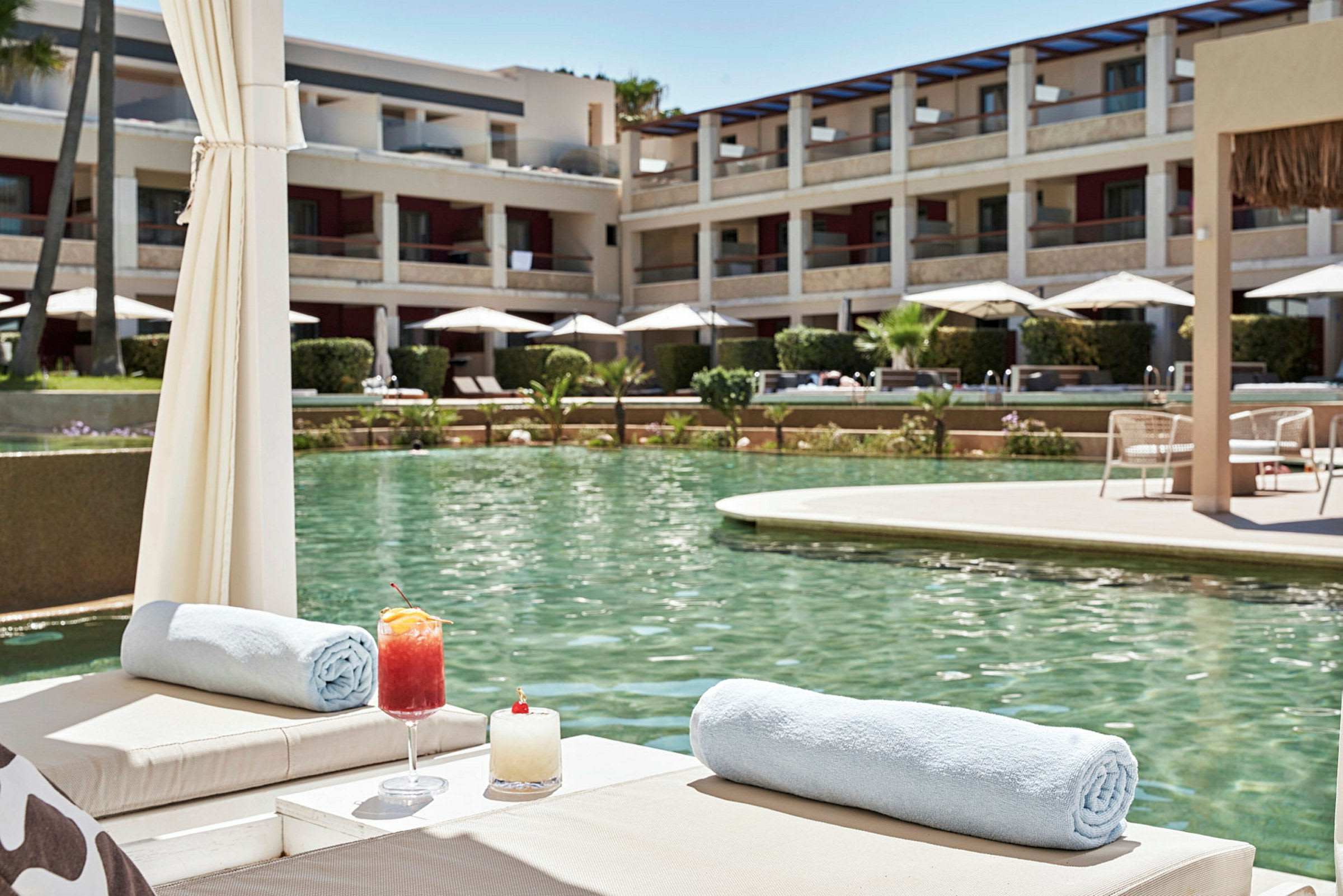 En pool med liggestole, drinks og håndklæder, omgivet af bygninger og parasoller. (AI-genereret tekst)
