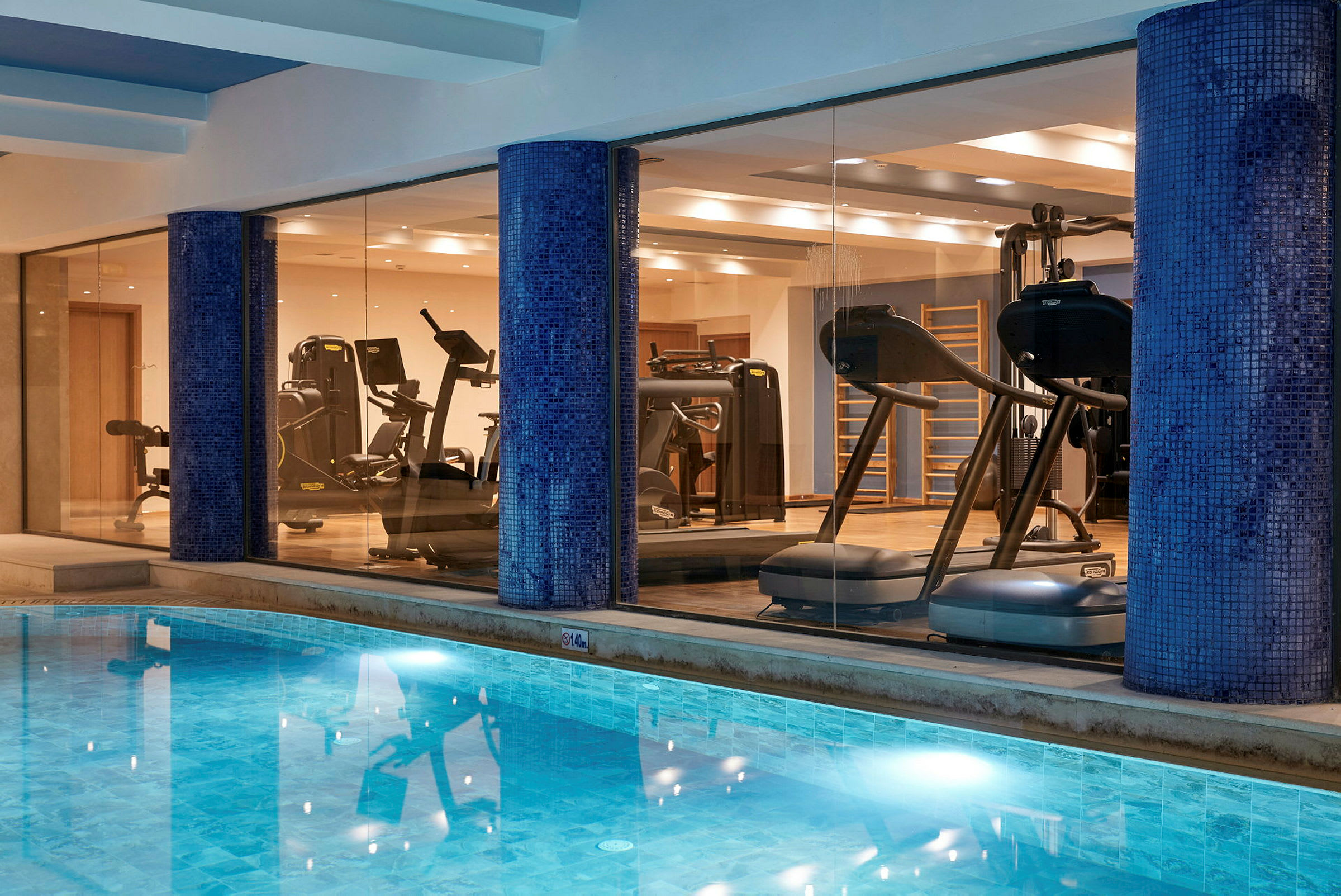 Indendørs pool med fitnessudstyr bag glasvægge. (AI-genereret tekst)