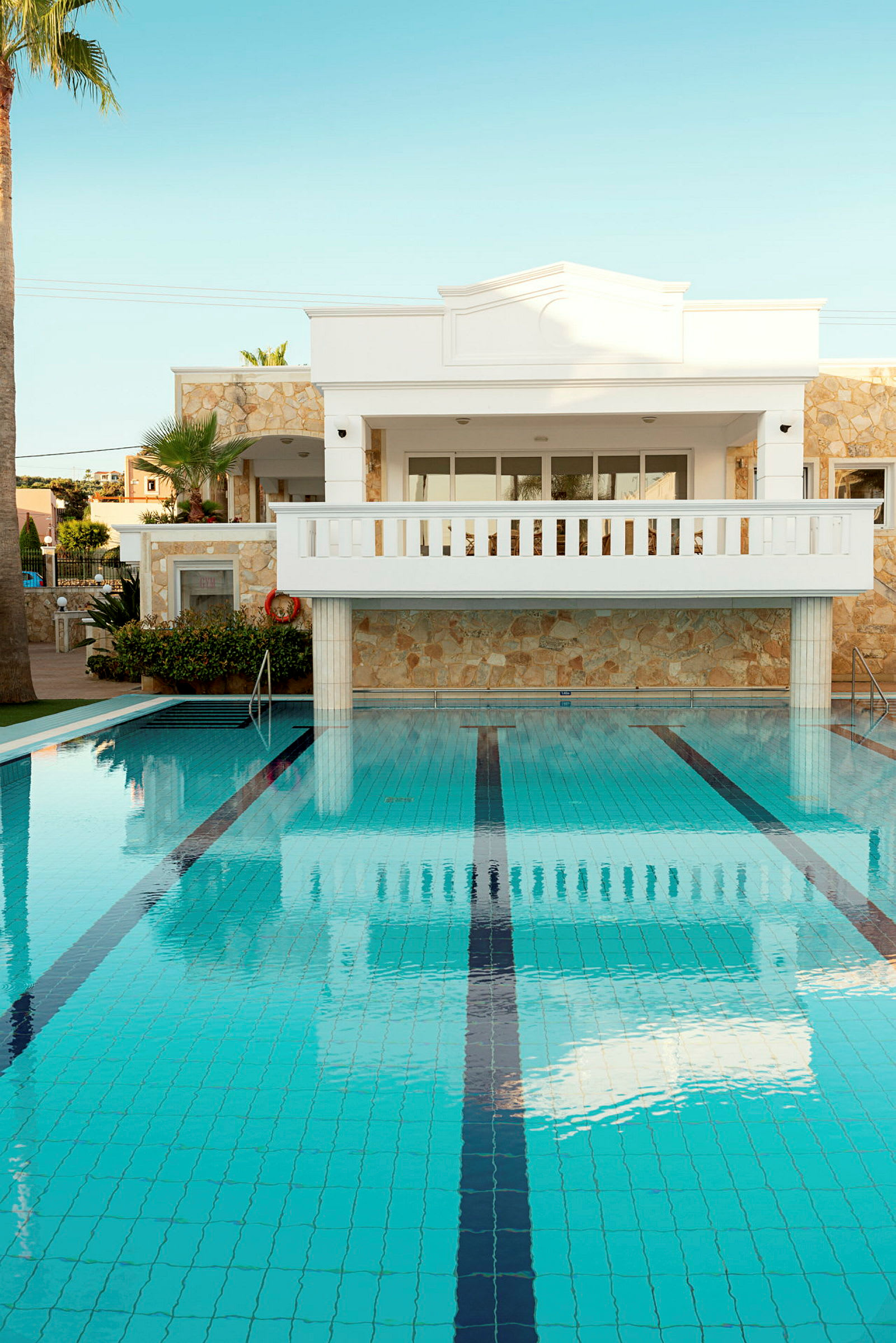 Hvid bygning med stenfacade og balkon foran en swimmingpool. (AI-genereret tekst)