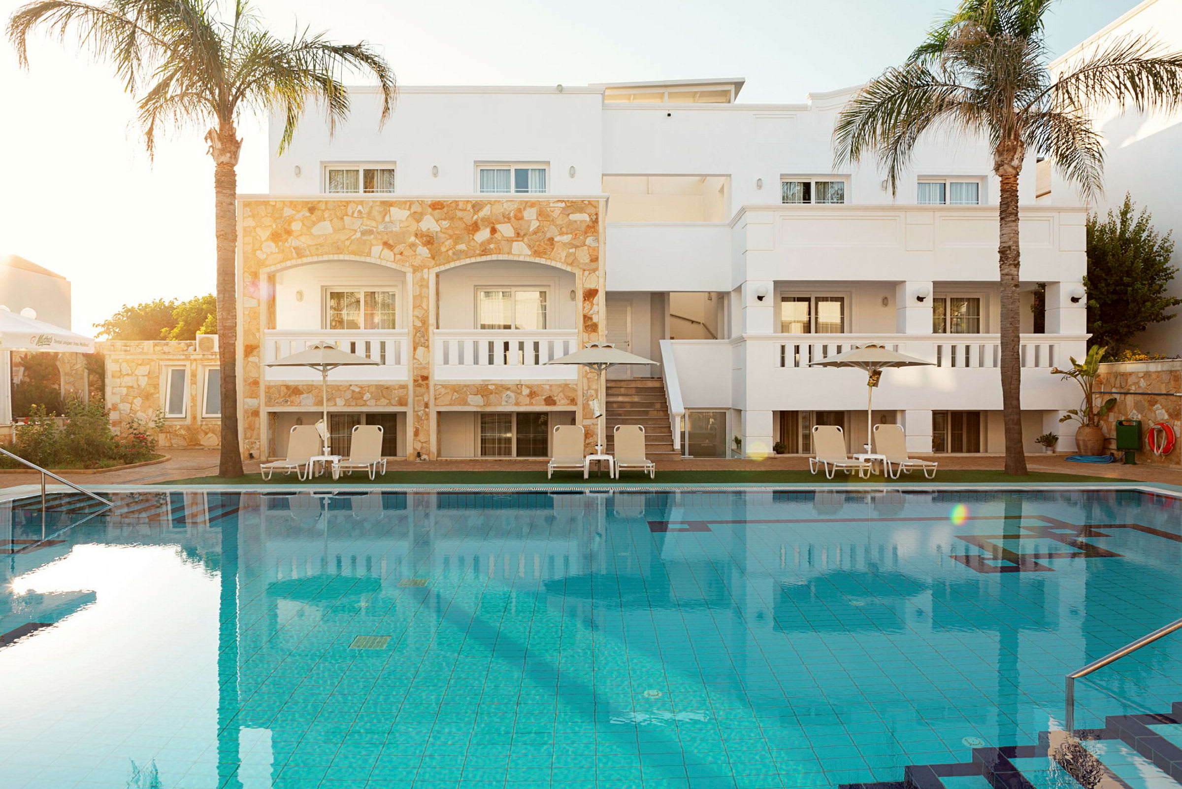 Billede af et hotelområde med en swimmingpool i forgrunden og liggestole under parasoller ved poolkanten foran en bygning. (AI-genereret tekst)