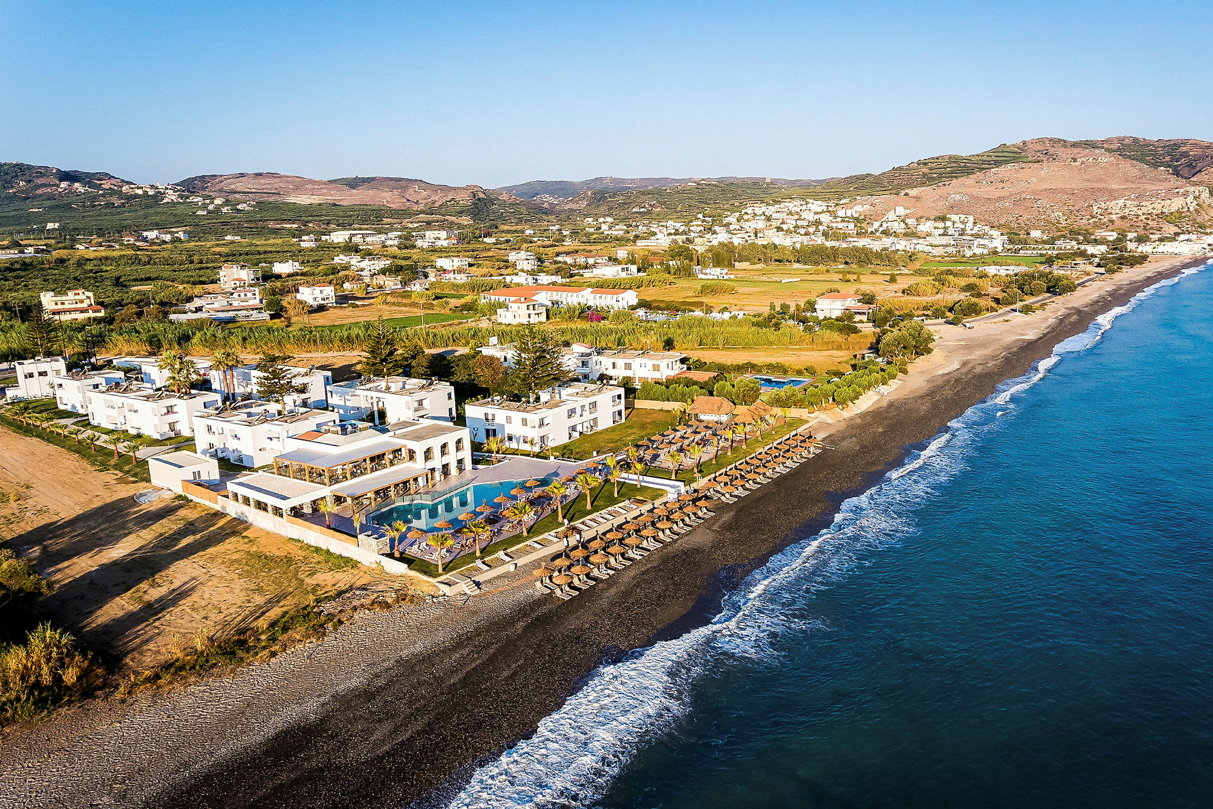 Udsigt over en strand med parasoller, liggestole og et resort med pools i nærheden. (AI-genereret tekst)