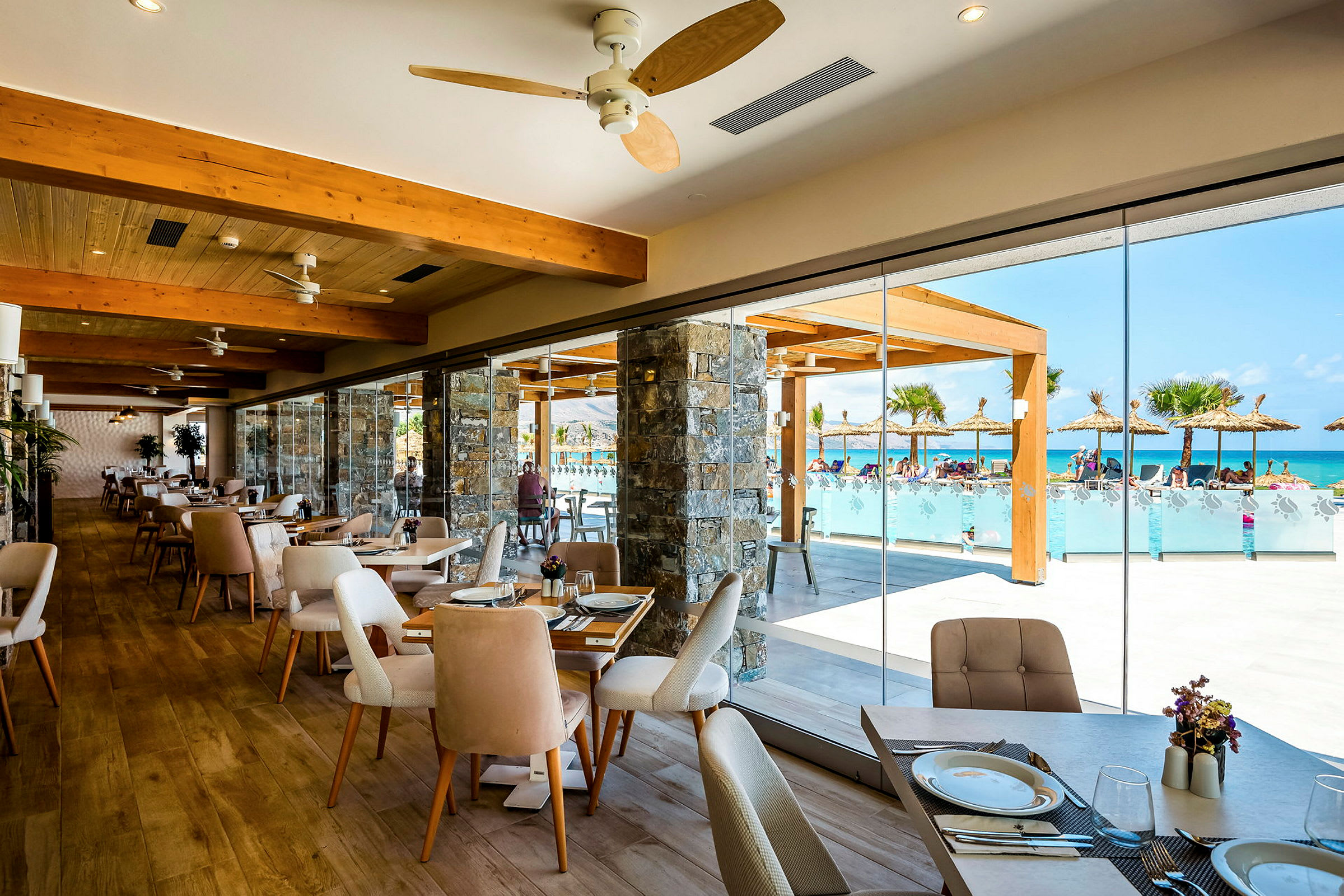 Restaurant med glasvægge, der vender mod en pool og palmebeklædte parasoller. (AI-genereret tekst)
