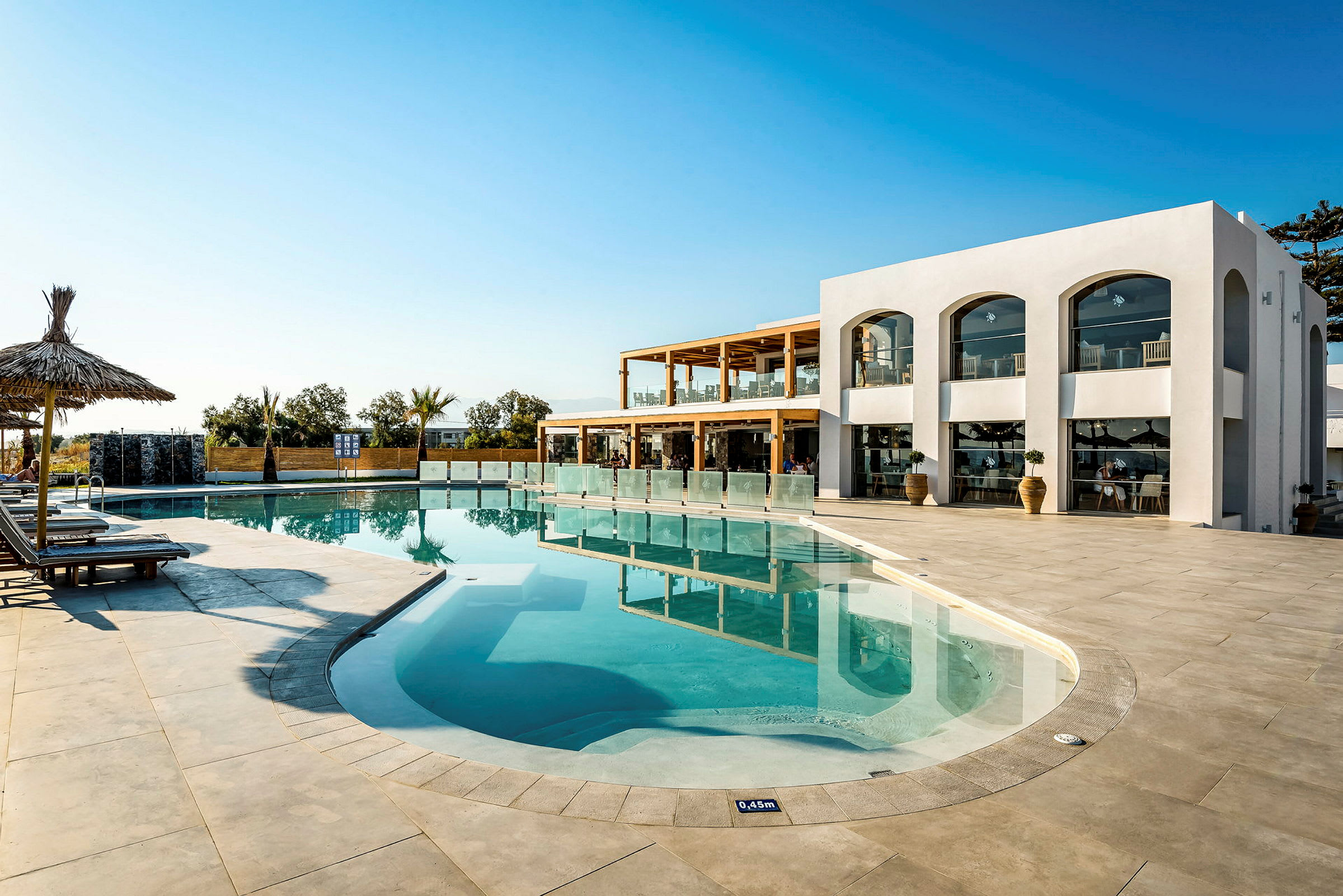 En udendørs pool ved et hotel med liggestole, parasoller og en moderne hvid bygning i baggrunden. (AI-genereret tekst)