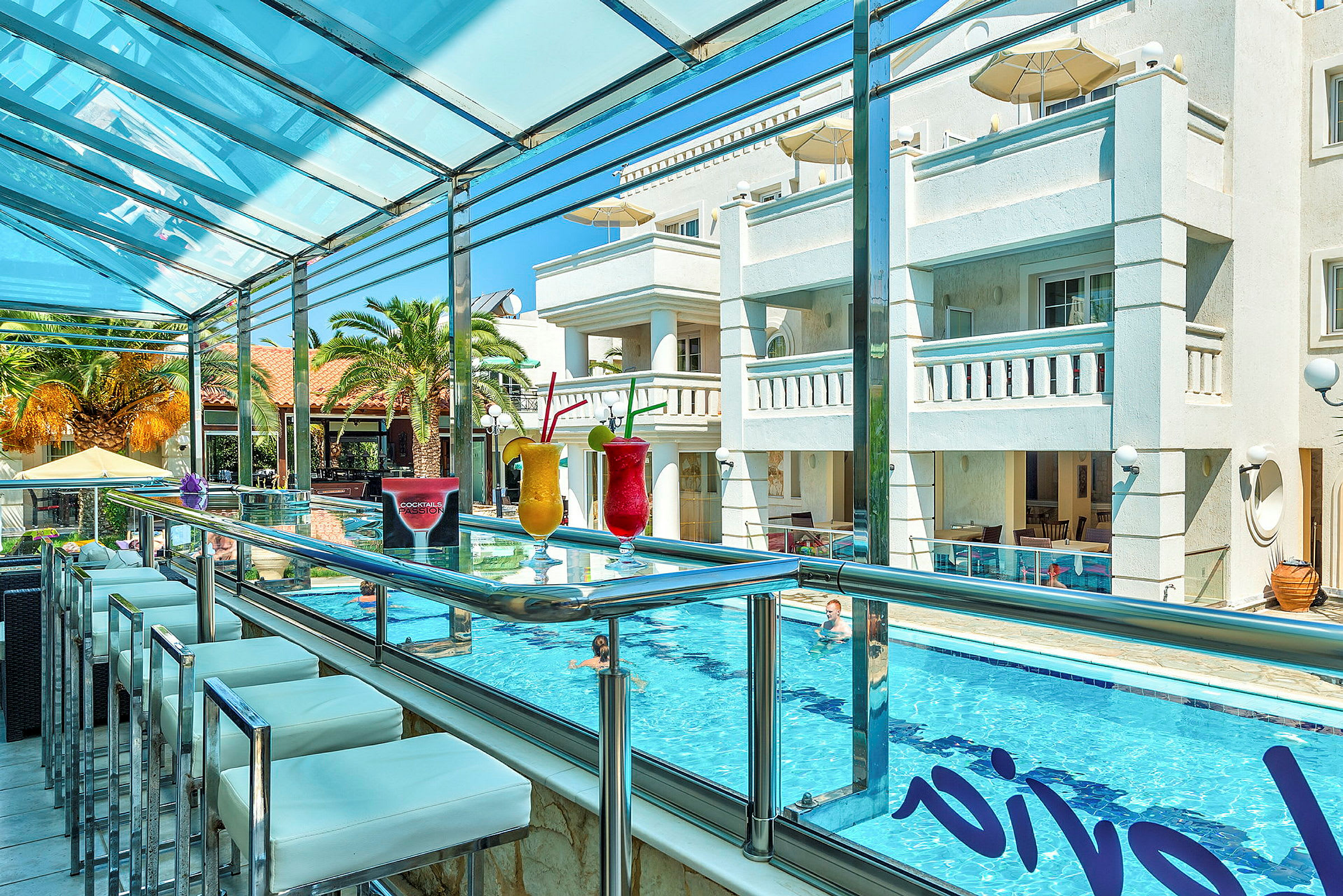 Billede af en udendørs bar med glasrækværk, hvor farverige cocktails er placeret på disken. I baggrunden ses en pool og en hotelbygning. (AI-genereret tekst)