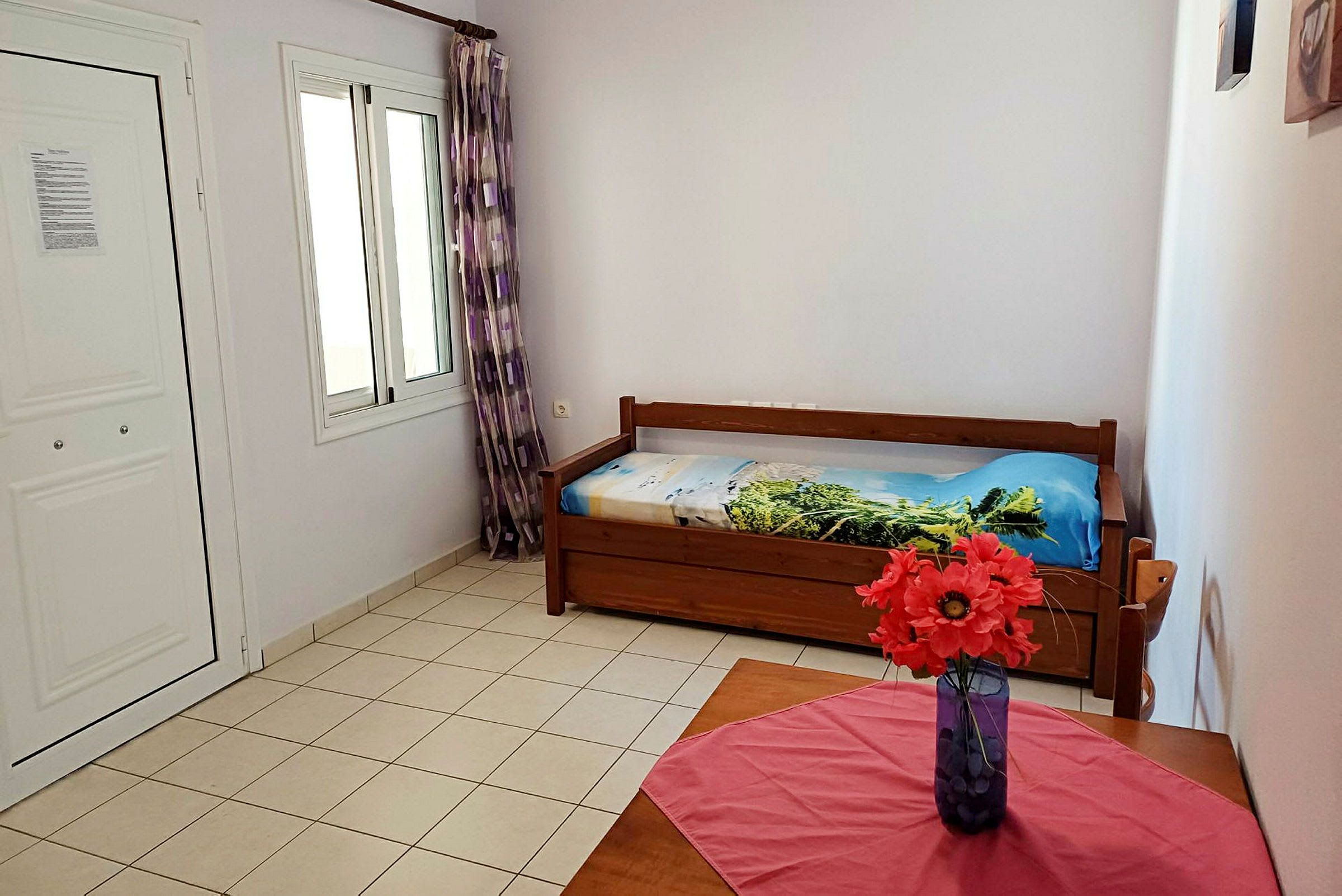 Et værelse med en enkeltseng og et bord med en vase med røde blomster. (AI-genereret tekst)