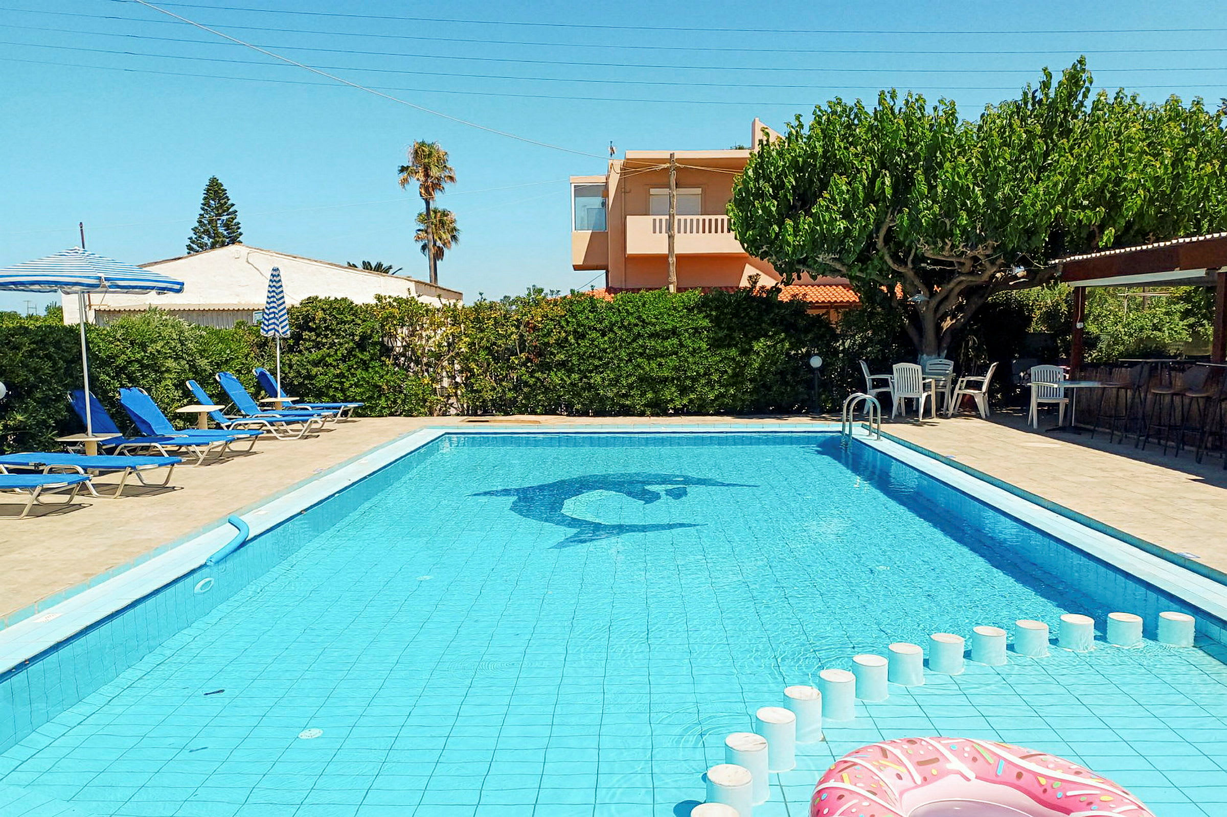 En udendørs pool med blå liggestole og parasoller på en solterrasse. (AI-genereret tekst)