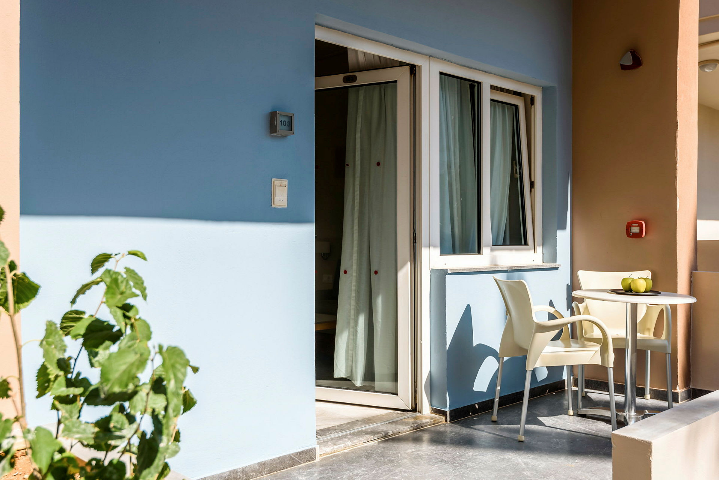 En balkon med to stole og et bord står foran en bygning med blå og beige vægge. (AI-genereret tekst)