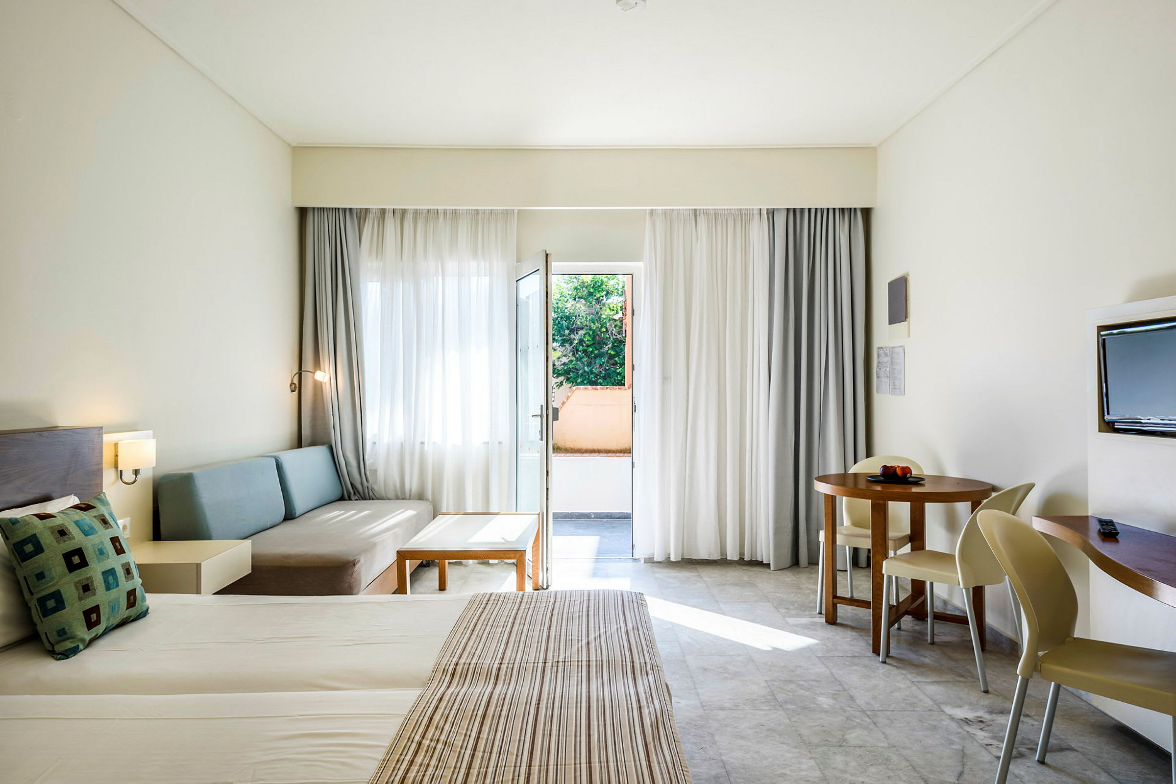 Hotelværelse med redt seng, siddeområde, rundt spisebord og altandør. (AI-genereret tekst)
