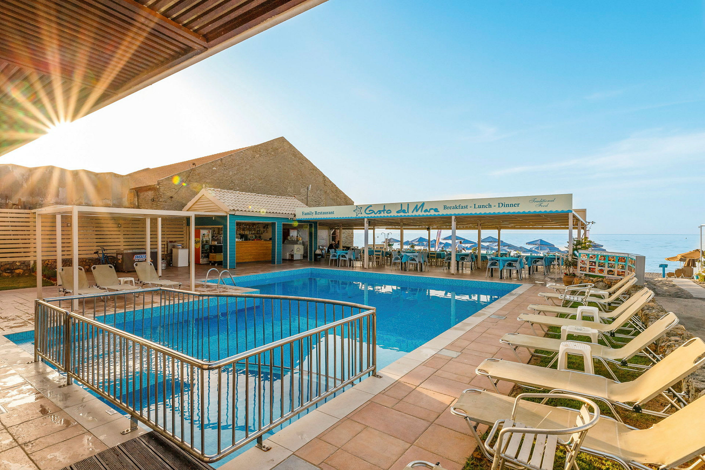 En udendørs swimmingpool med liggestole omkring og en restaurant i baggrunden ved kysten. (AI-genereret tekst)