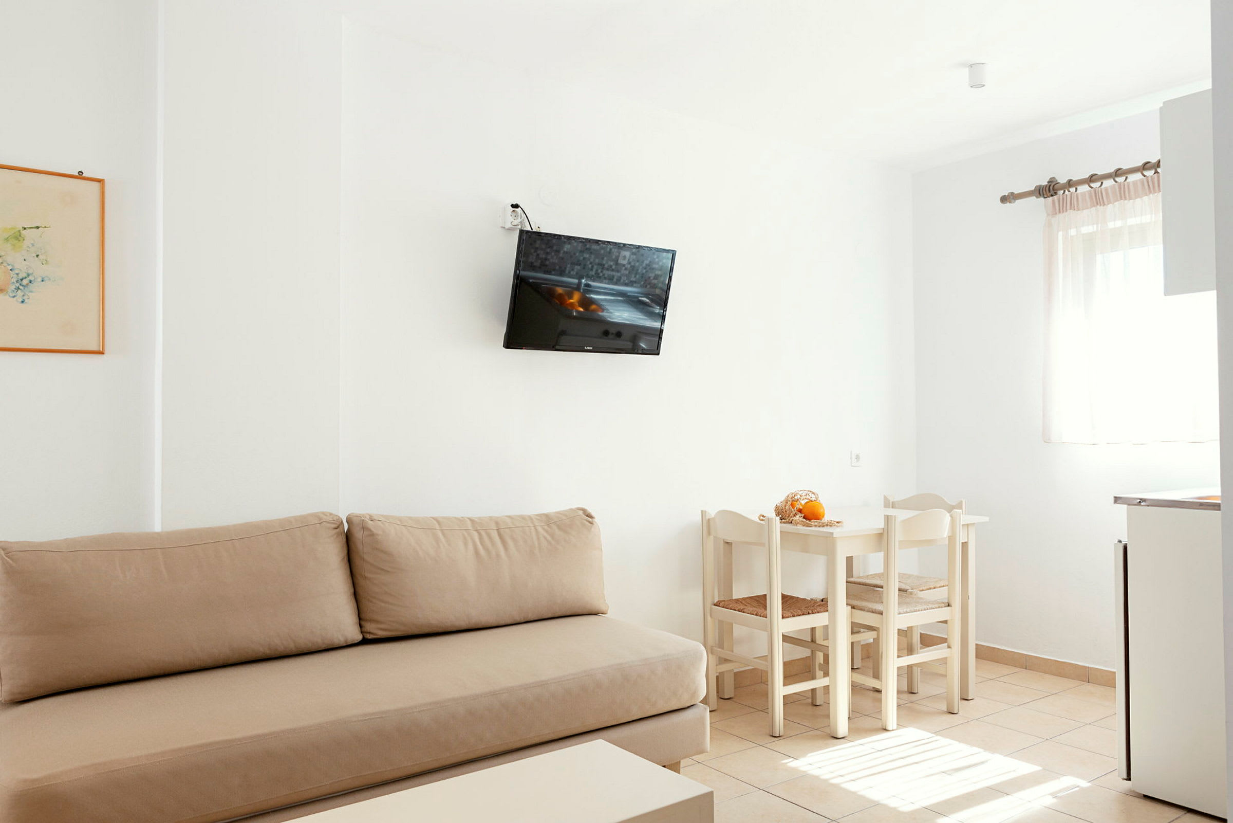 Et lyst rum med beige sofa, væghængt TV, hvidt spisebord med stole og et vindue med lyse gardiner. (AI-genereret tekst)