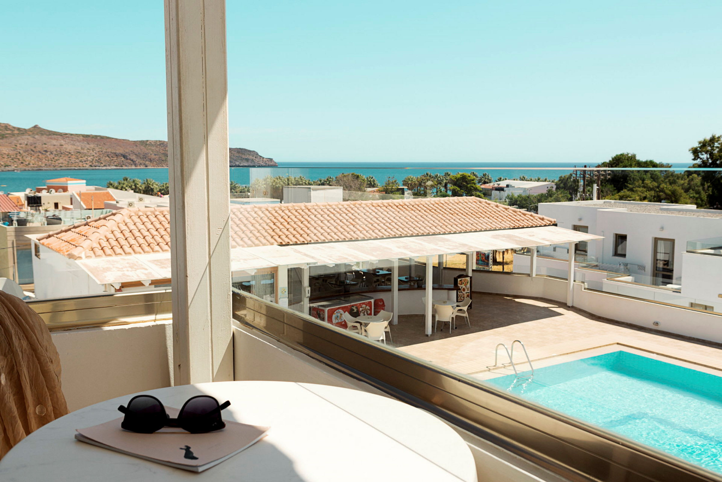 Udsigt fra en balkon over en pool, dele af hotelområdet og havet i baggrunden. (AI-genereret tekst)