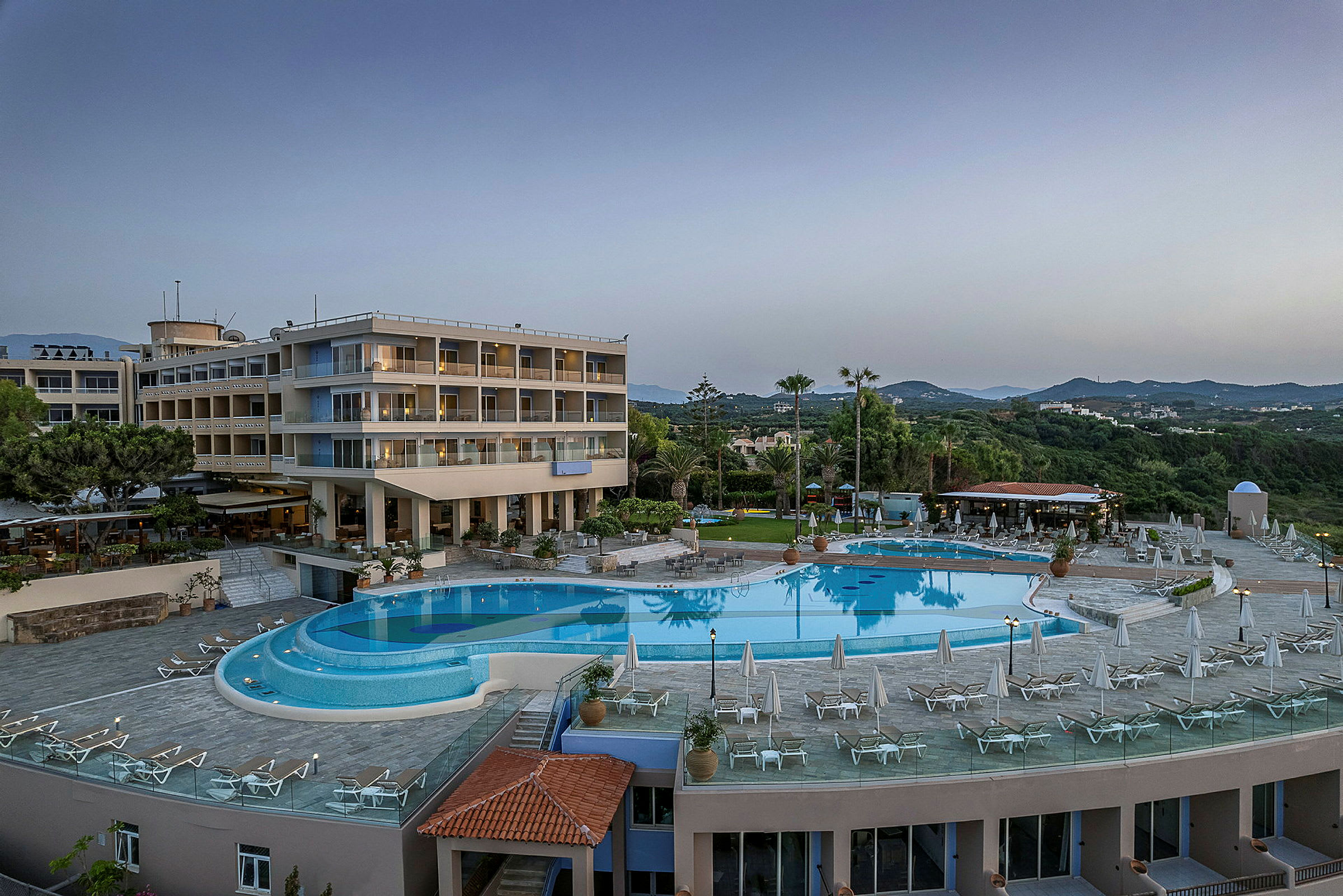 Hotel med flere etager, swimmingpool og liggestole i grønne omgivelser. (AI-genereret tekst)