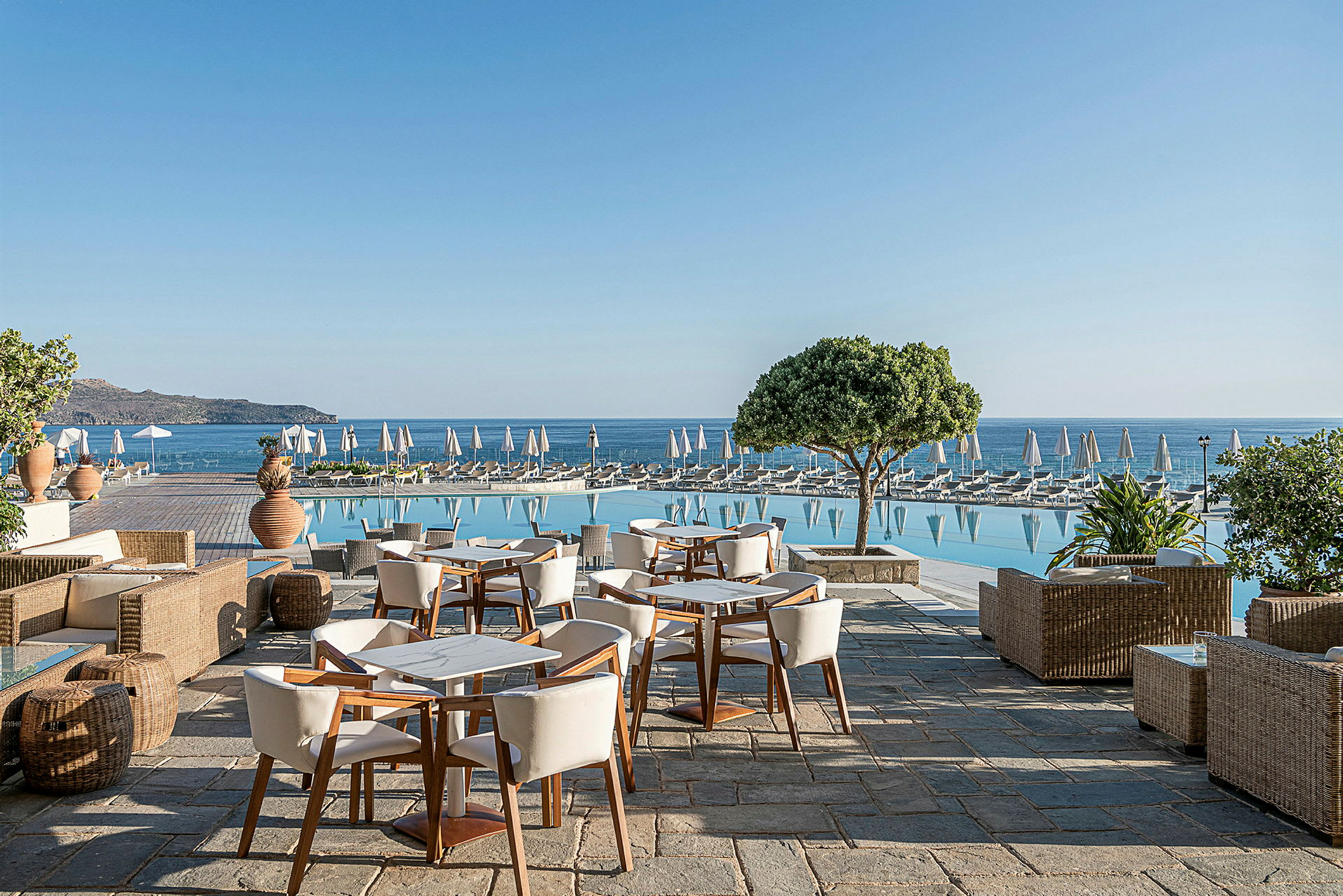 Udendørs terrasse med borde og stole, en pool med liggestole ved siden af, og havet i baggrunden. (AI-genereret tekst)