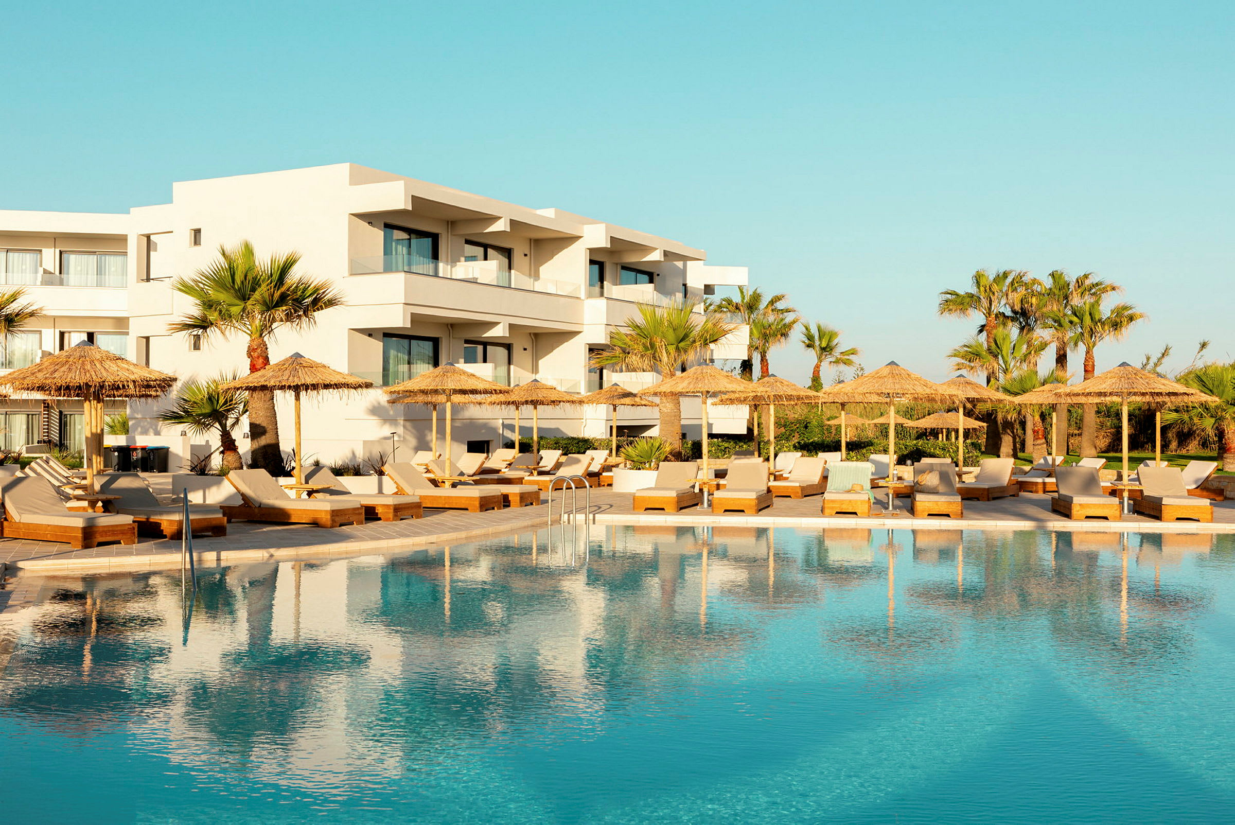 Hotel med hvid facade bag en swimmingpool og liggestole med parasoller. (AI-genereret tekst)
