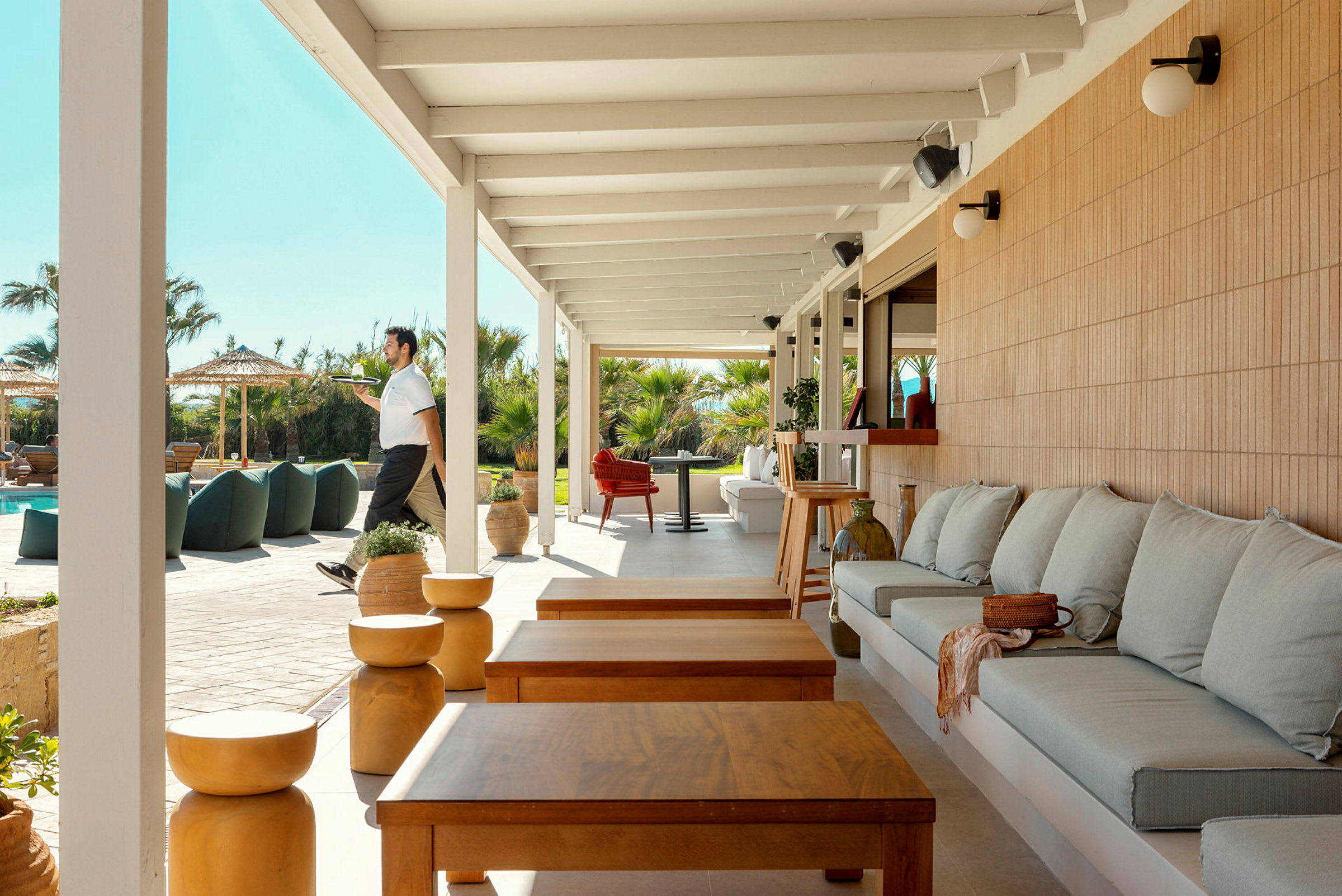 En terrasse med siddepladser ved siden af en udendørs pool, omgivet af palmer og parasoller. (AI-genereret tekst)