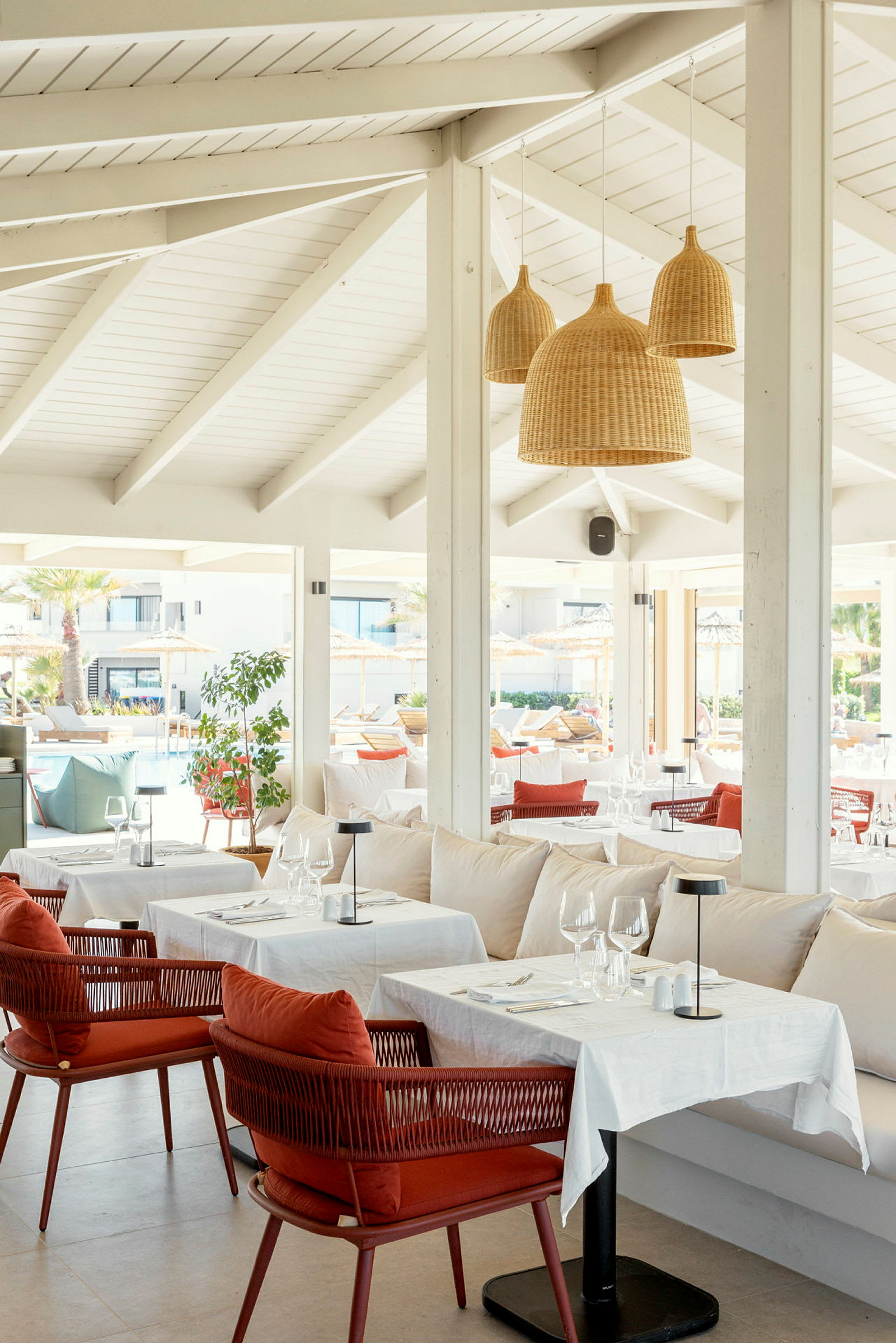 Interiør af en restaurant med hvide loftsbjælker, hængende fletlamper samt dækkede borde og stole. (AI-genereret tekst)
