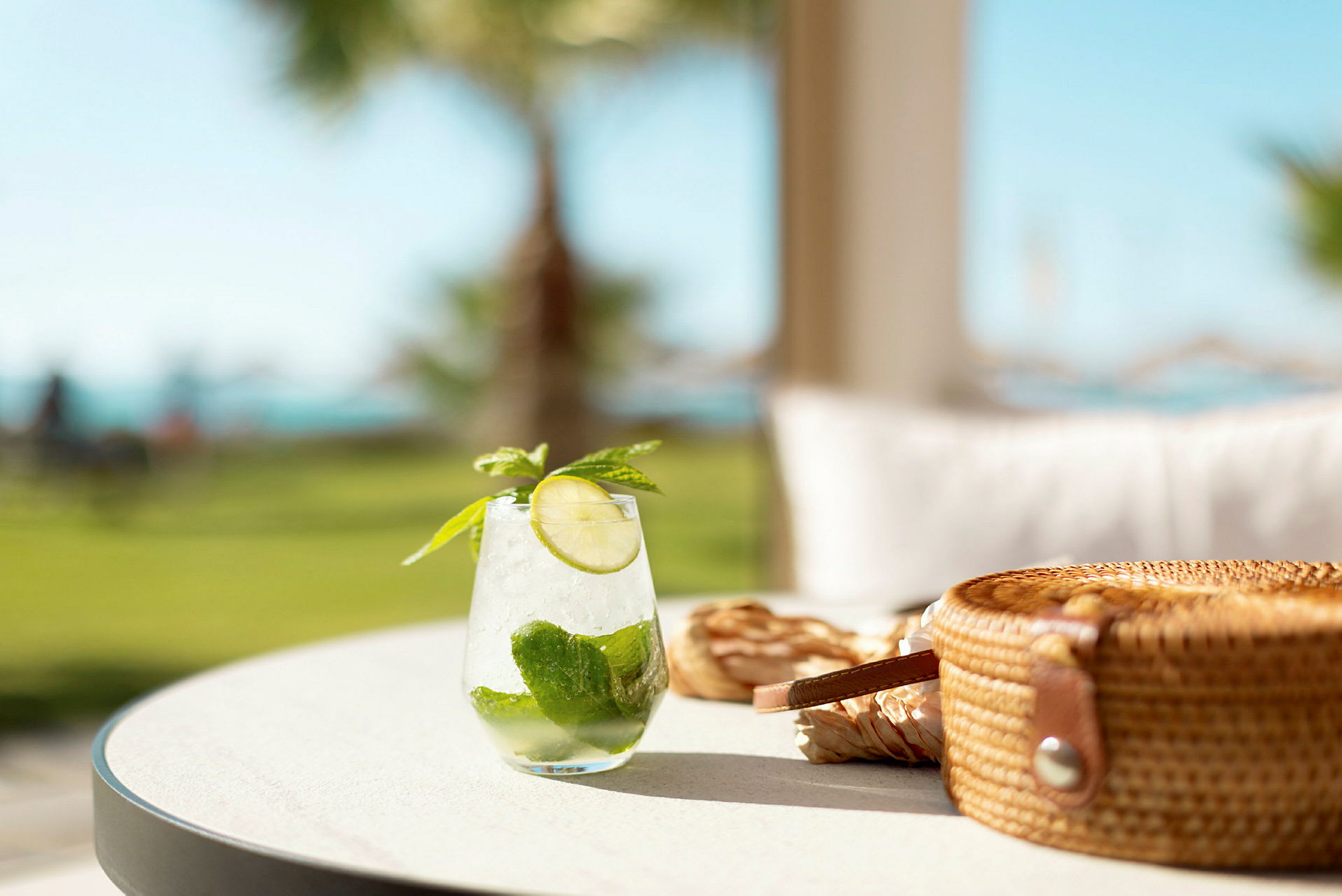 Glas med kold drink med lime og mynte på et bord, med en stråtaske ved siden af. (AI-genereret tekst)