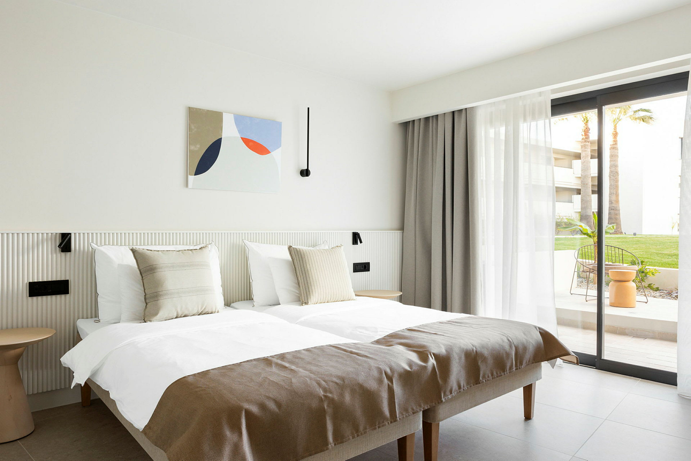 Et lyst hotelværelse med to enkeltsenge, neutrale detaljer og en glasdør ud til en terrasse. (AI-genereret tekst)